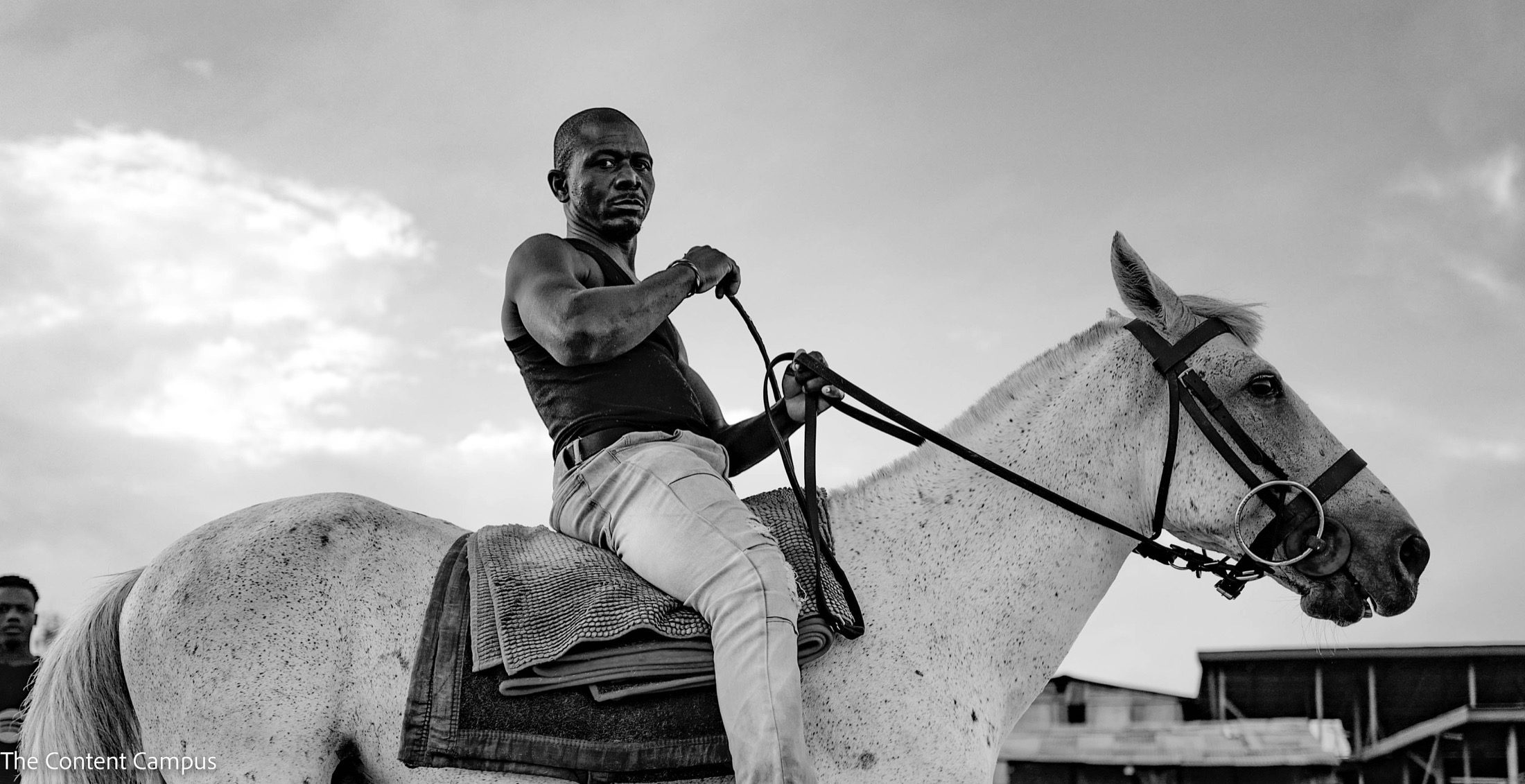 Black-and-white photo of a sleeveless man riding a speckled white horse, holding the reins and looking toward the camera under a cloudy sky, with low buildings behind.