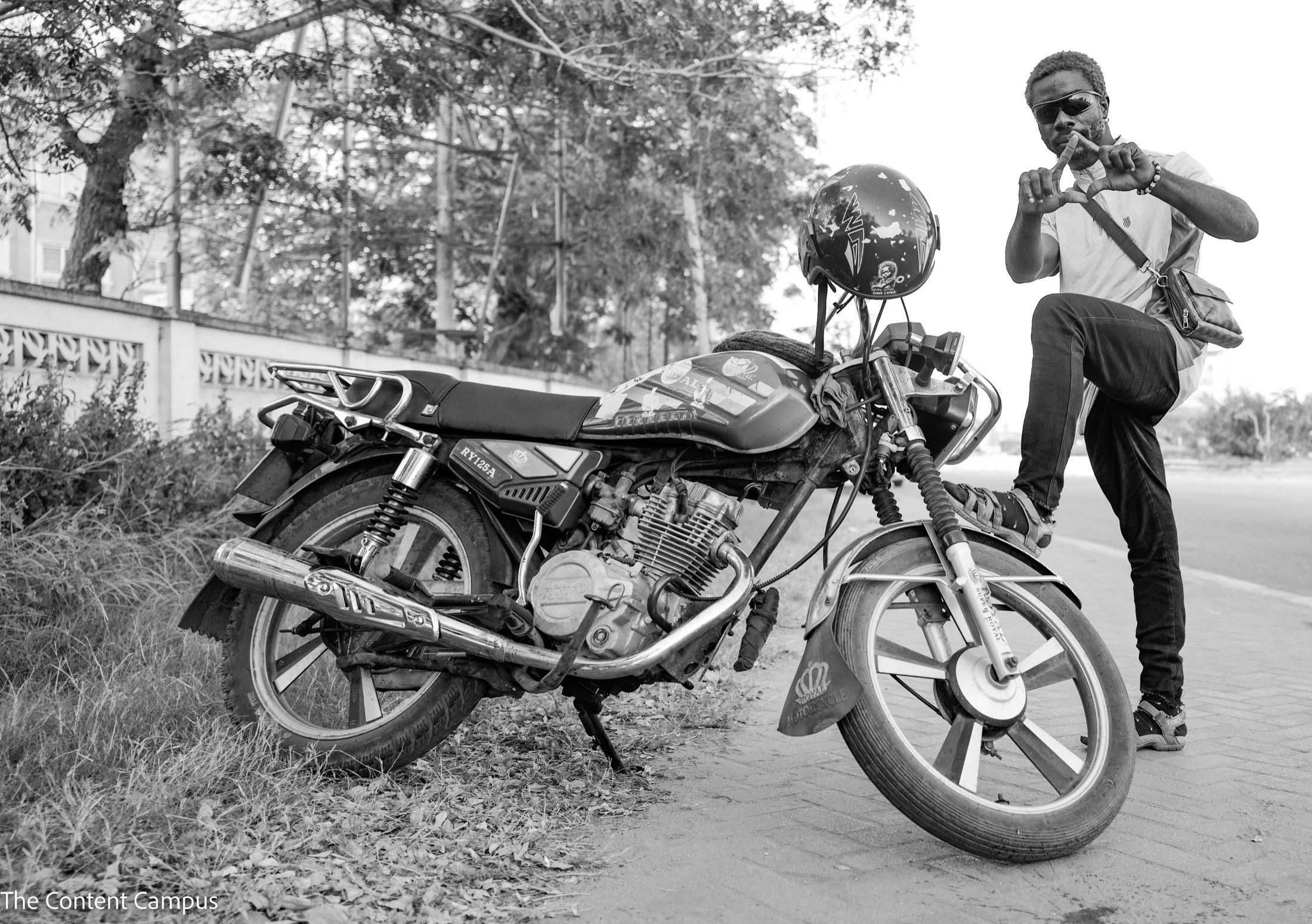 Black-and-white image of a man in sunglasses with one foot raised on a parked motorcycle, forming a triangle with his hands, beside a tree-lined wall and paved road.