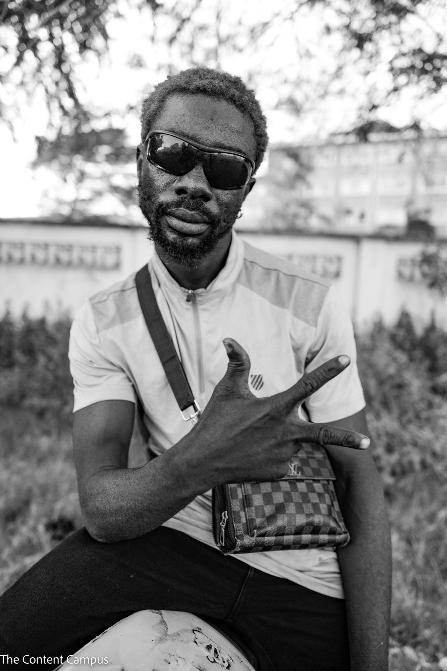 Black-and-white close portrait of a bearded man in sunglasses and a zip-neck shirt, holding up a hand gesture over a checkered shoulder bag, with a blurred wall and foliage behind.