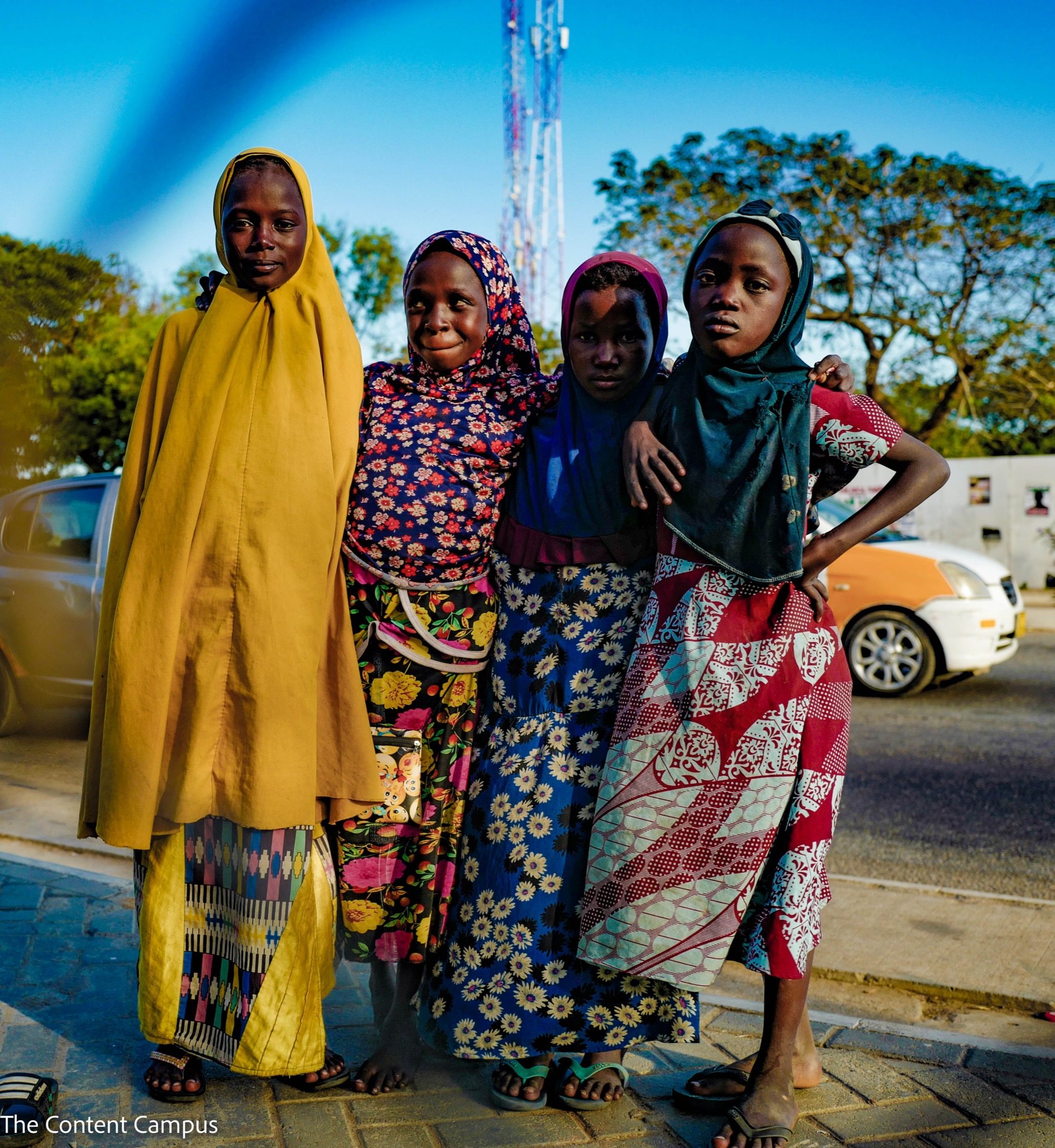 Four young girls in colorful patterned dresses and headscarves stand arm in arm on a paved sidewalk in warm light, with a cell tower, trees and cars behind.