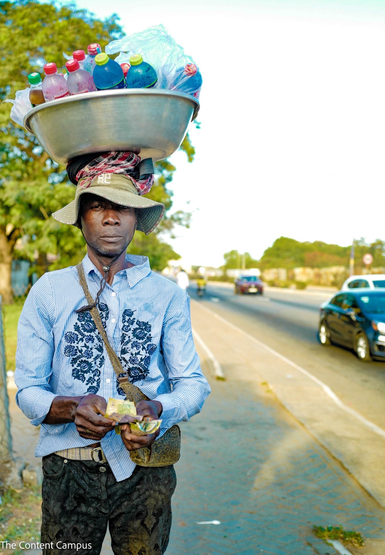 A street vendor in a bucket hat balances a large metal basin of bottled drinks on his head, holding banknotes beside a road with passing cars.