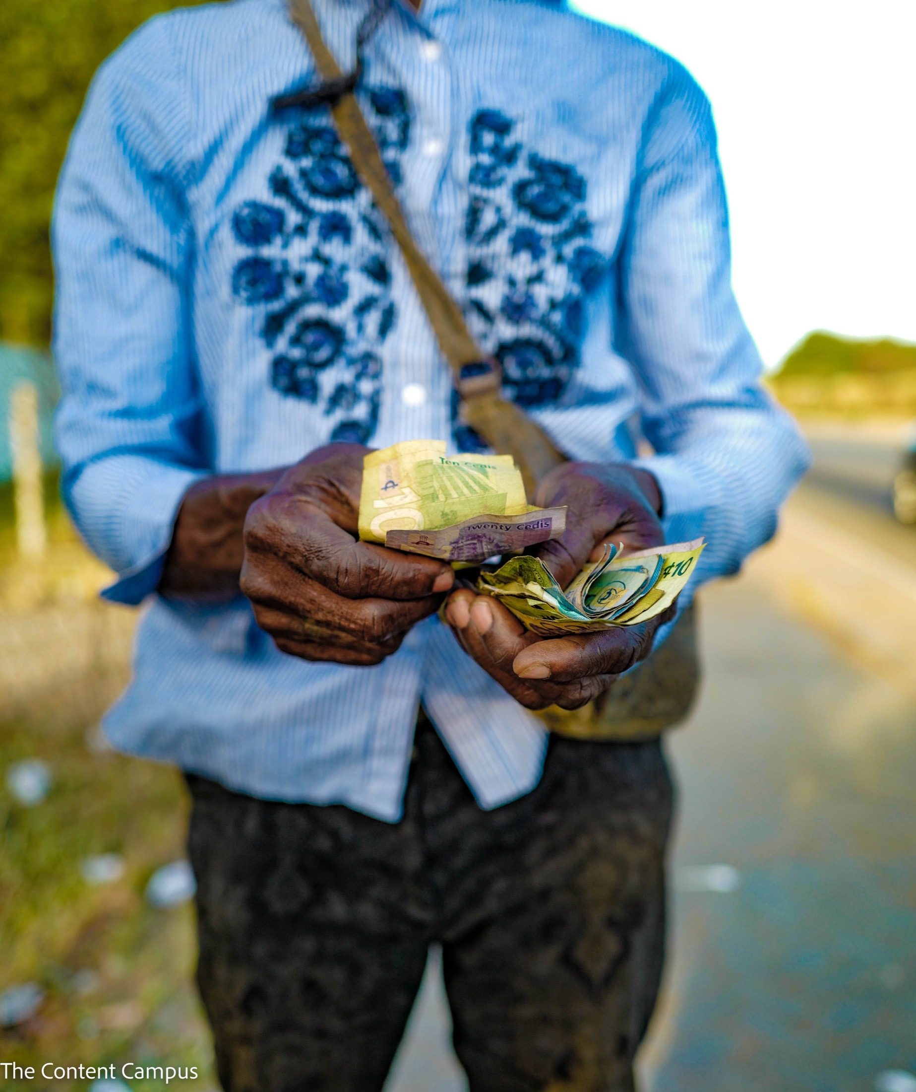 Close view of a person's hands holding worn Ghanaian cedi banknotes, wearing a blue striped shirt with dark floral embroidery and a strap across the chest, with the road blurred behind.