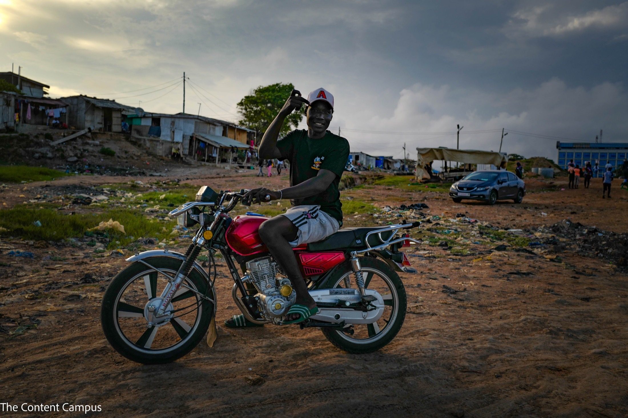 A smiling man in a cap and green T-shirt sits on a red motorcycle, one hand raised to his head, on dirt ground under stormy dusk skies.