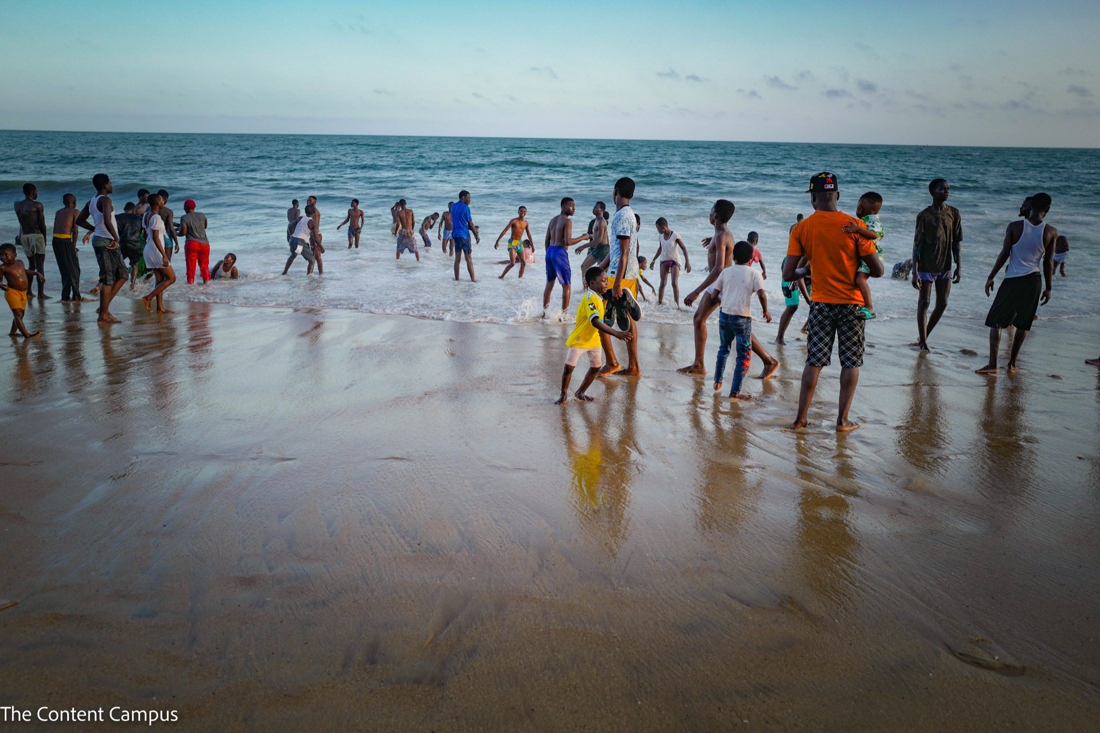 Dozens of people, including children, wade and play in shallow surf on a wet sandy beach beneath a pale blue evening sky.