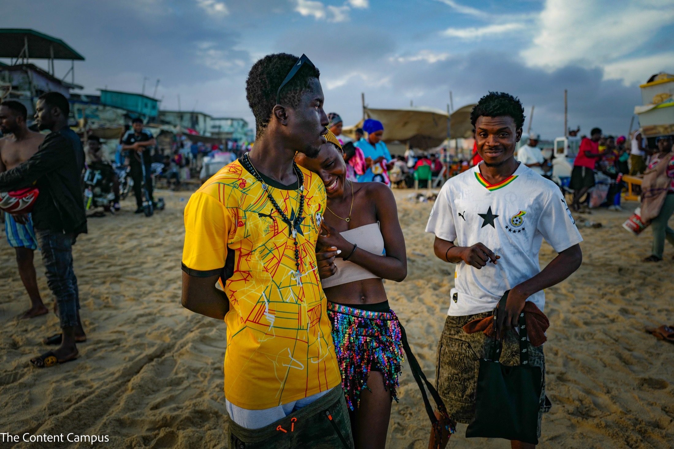 Two young men in yellow and white football shirts stand on a crowded beach with a woman leaning against one's shoulder, market stalls behind.