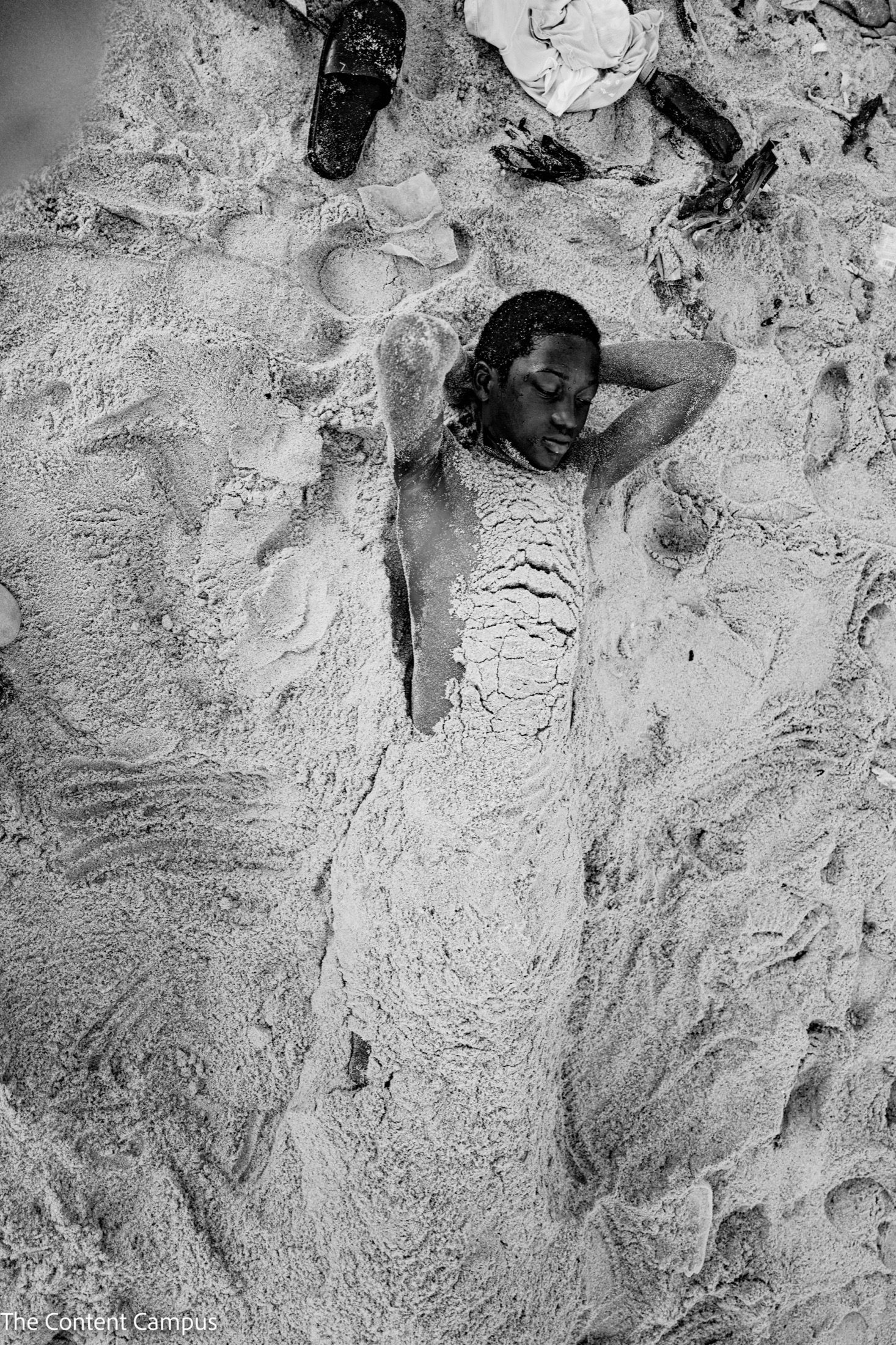 Seen from above in black and white, a person lies on their back with hands behind their head, body covered in sand, a sandal nearby.