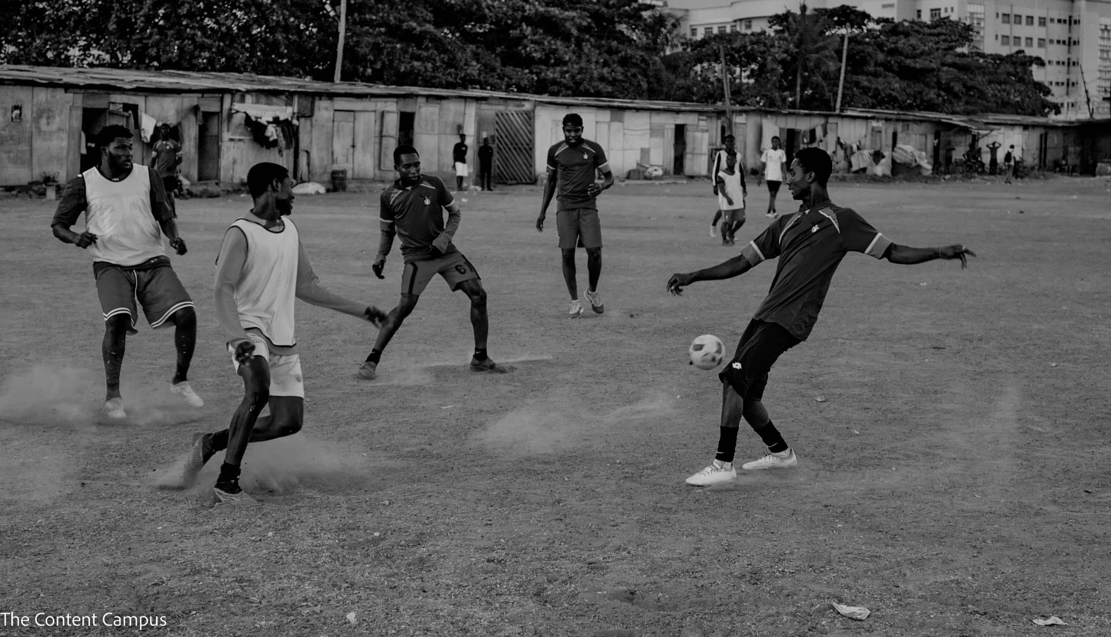 In black and white, men in bibs and dark shirts contest a football on a dusty pitch, with low shacks and trees behind.