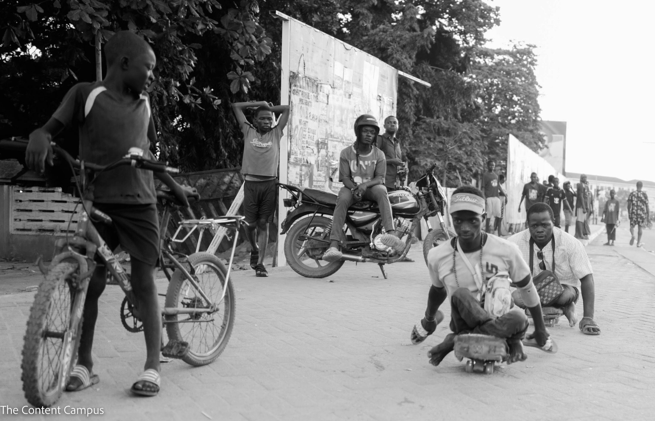Black-and-white street scene: two men ride low wheeled boards along a paved road while a boy on a bicycle, a seated motorcyclist and other onlookers watch.