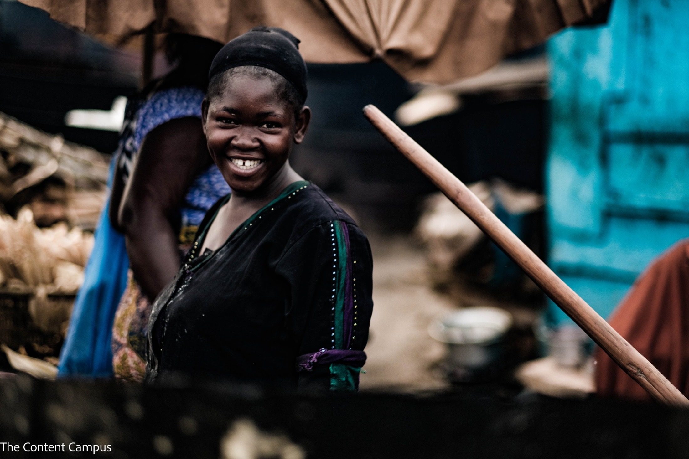 A woman in a dark top and black head covering smiles over her shoulder at a market stall, with a wooden pole and blurred blue wall behind.