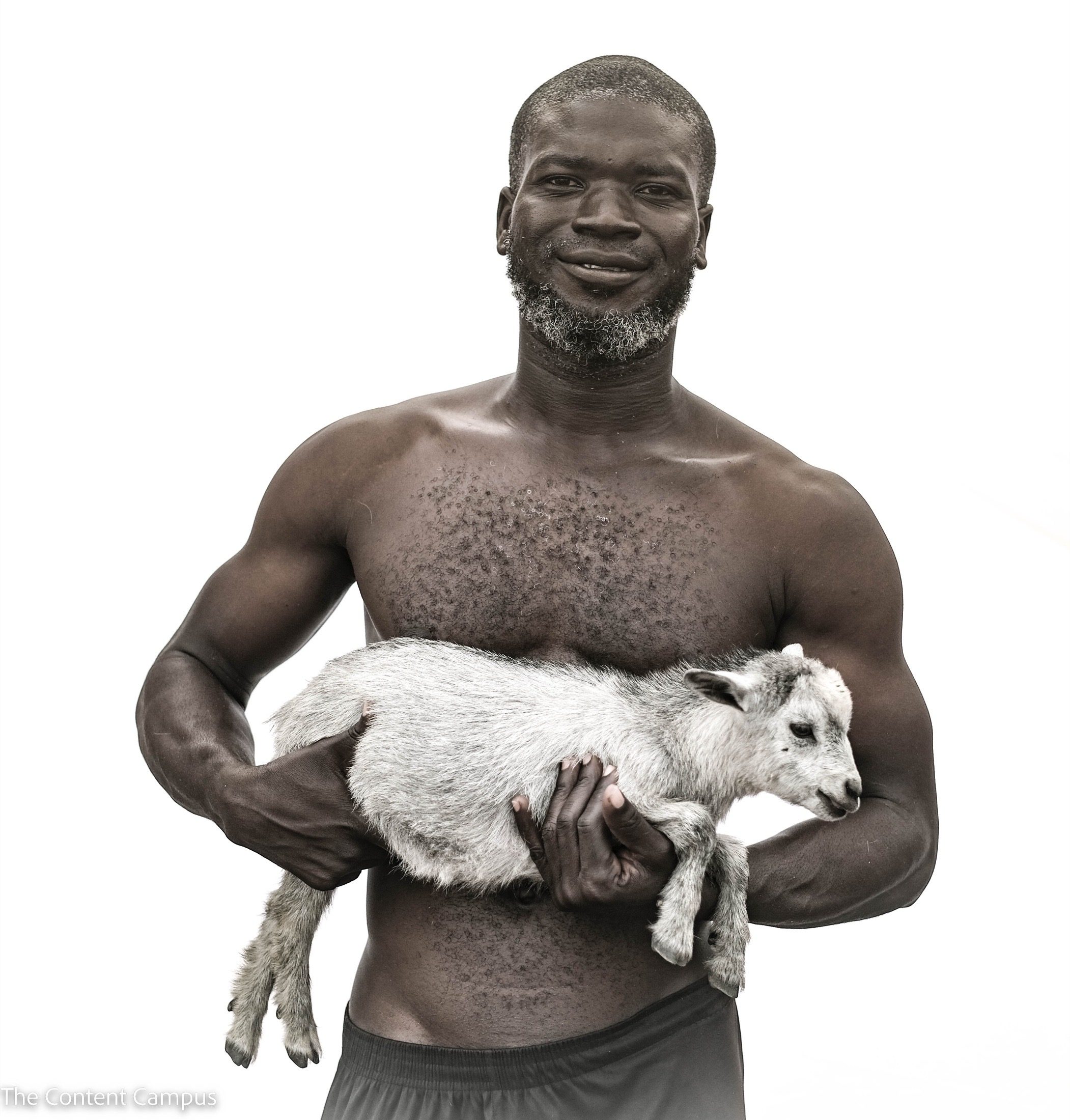 A shirtless, bearded man stands against a plain white background, holding a small pale goat horizontally in his arms and looking at the camera.