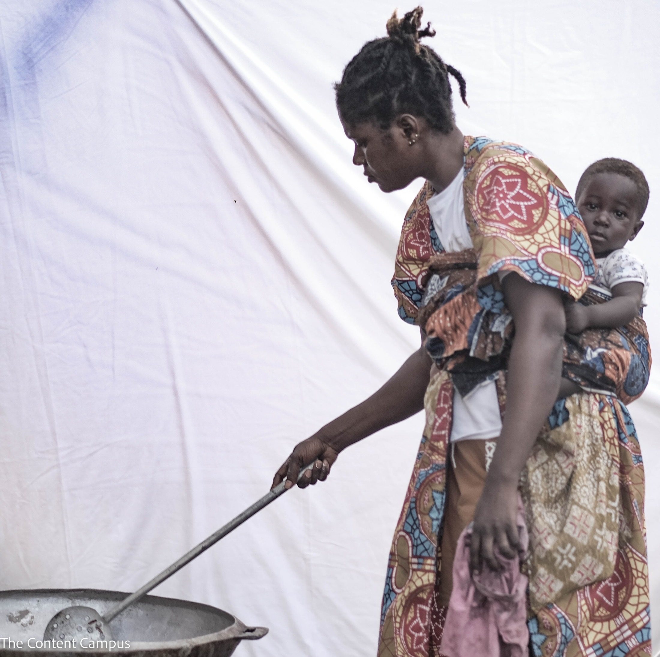 A woman in a colorful patterned dress carries a child on her back in a wrap while stirring a large pot with a long ladle.