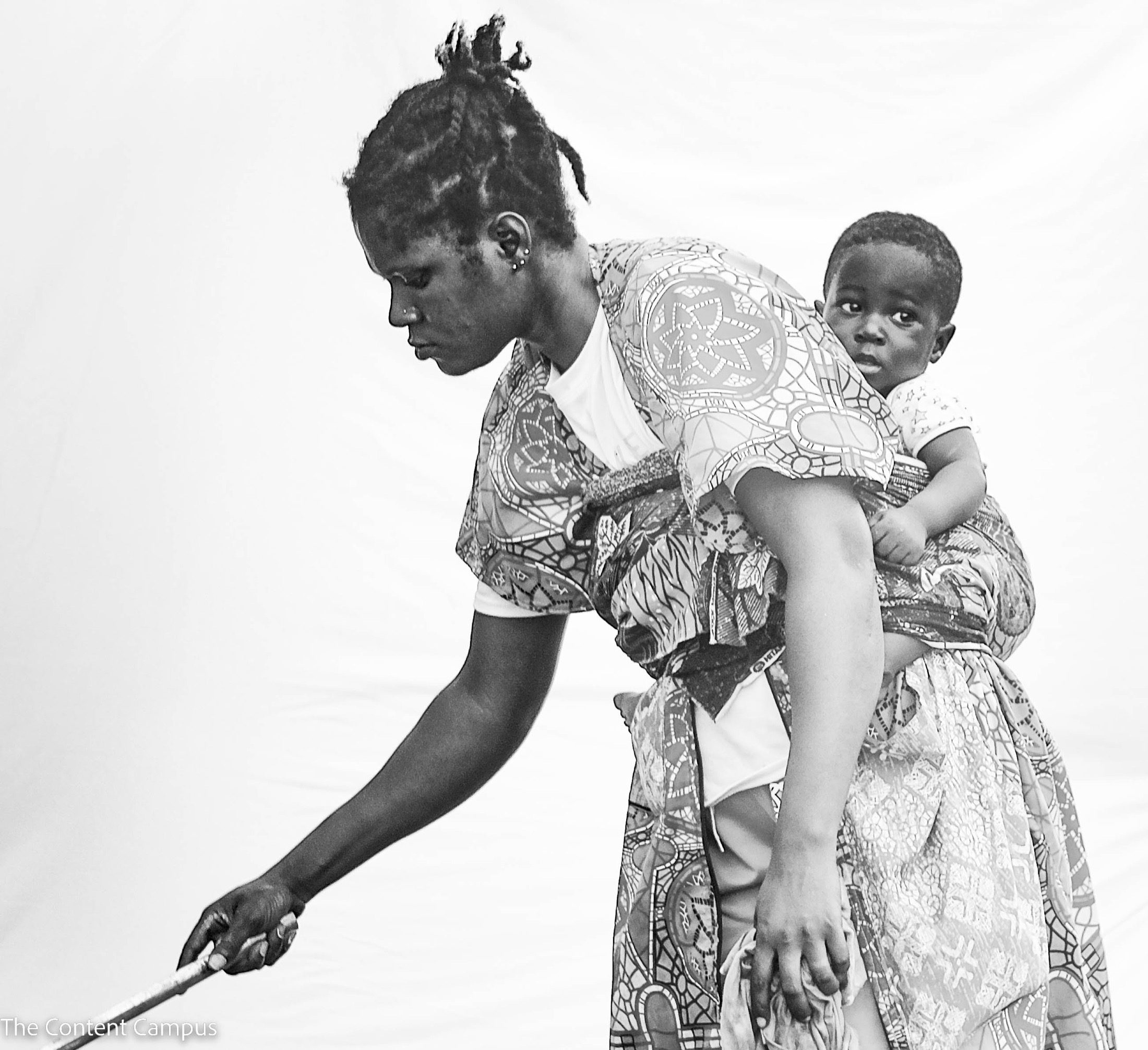 In black and white, a woman with braided hair bends forward holding a stick, a child in a wrap looking from her back.