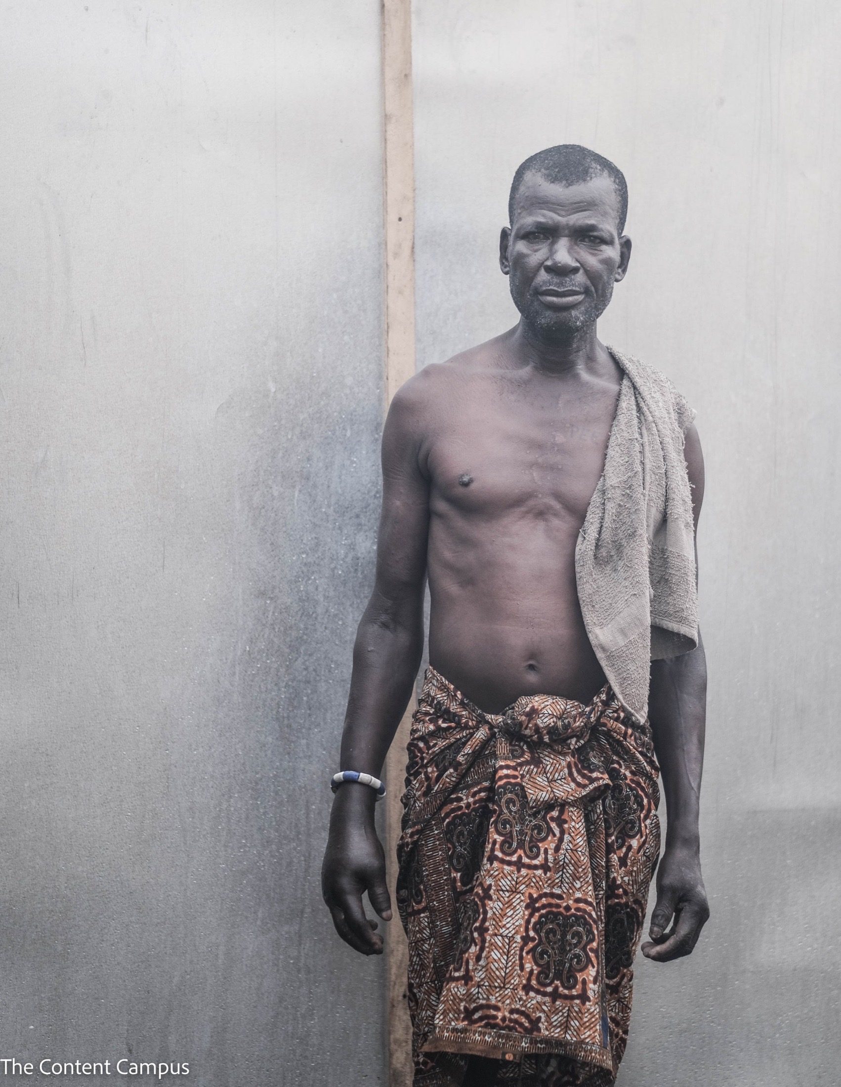 A shirtless man in a patterned wrap skirt, a towel over one shoulder and a beaded bracelet, stands before a pale, weathered wall.