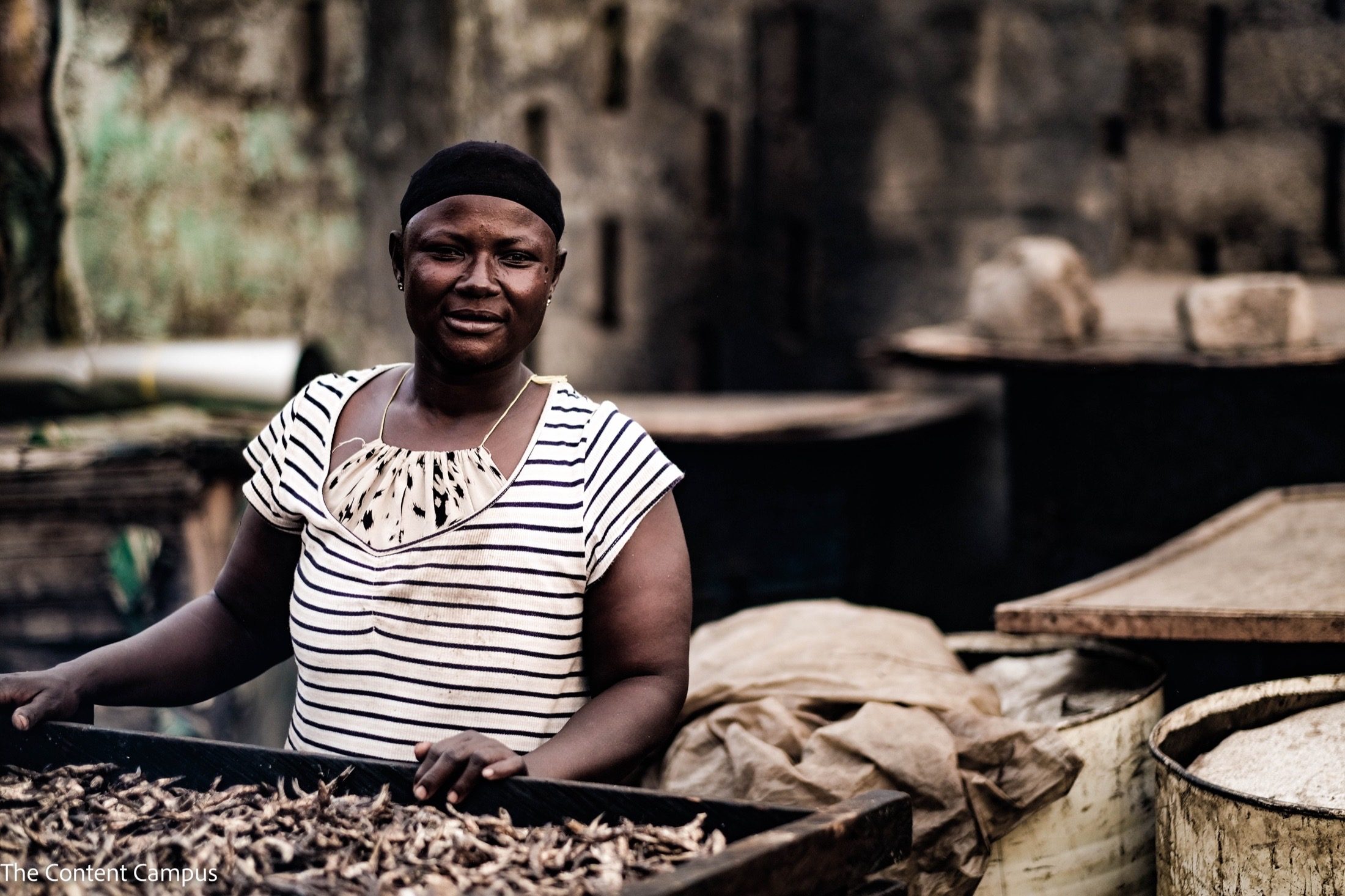 A woman in a striped top and dark cap stands behind a wooden tray of small dried fish, with large drums and a weathered wall behind her.