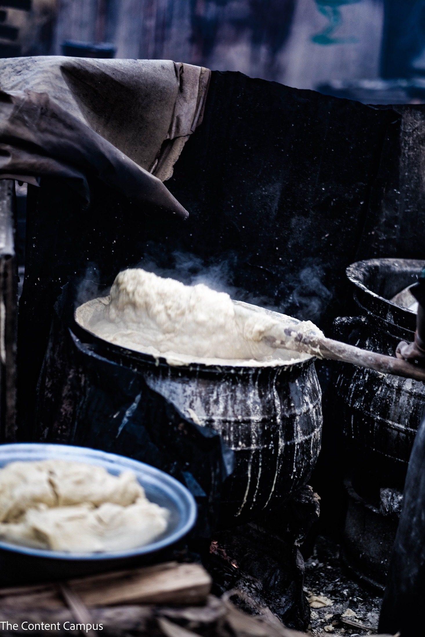 Steam rises from a large dark pot of thick pale paste being stirred with a long wooden paddle, with a bowl of the paste in the foreground.