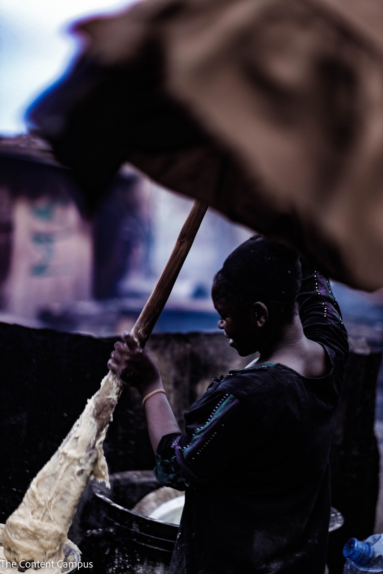 In silhouette, a person in a dark top stirs thick pale paste with a long wooden stick, while a blurred brown cloth arcs across the frame.