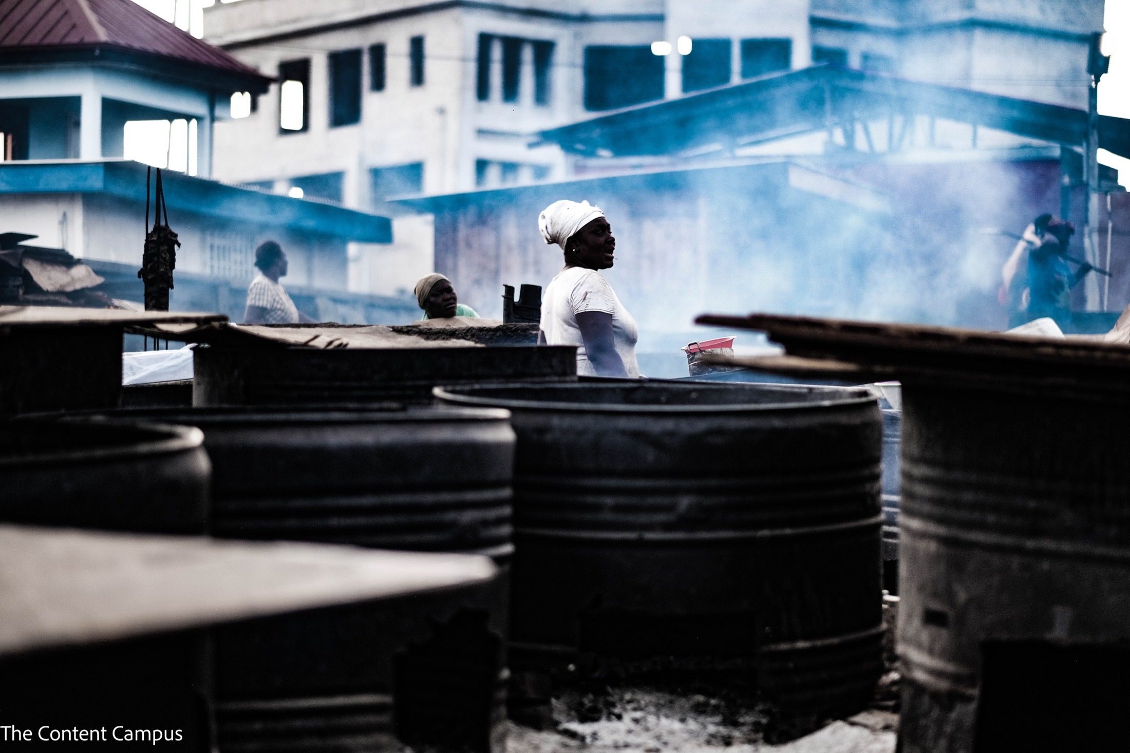 A woman in a white headwrap and light top stands among rows of dark metal drums, with smoke drifting past other workers and pale buildings behind.