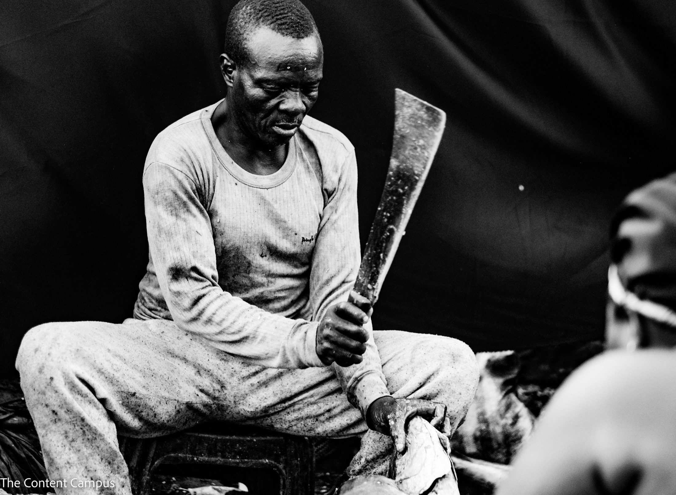 In black and white, a seated man in a worn, stained long-sleeved shirt holds a large machete upright while gripping a fish, against a dark backdrop.