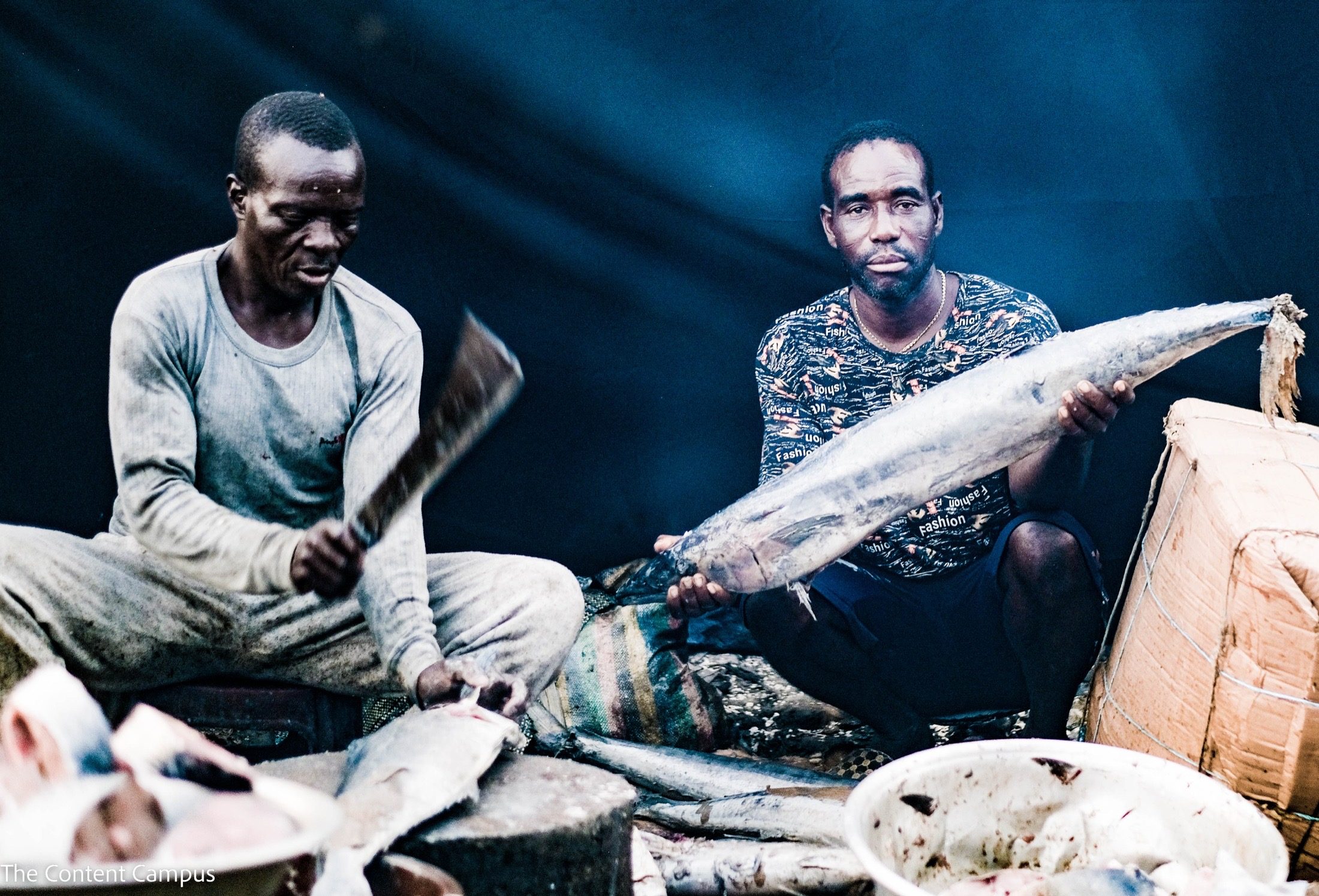 Two men sit among fish at a market stall under a dark blue tarp: one chops with a cleaver, the other holds a large fish toward the camera.
