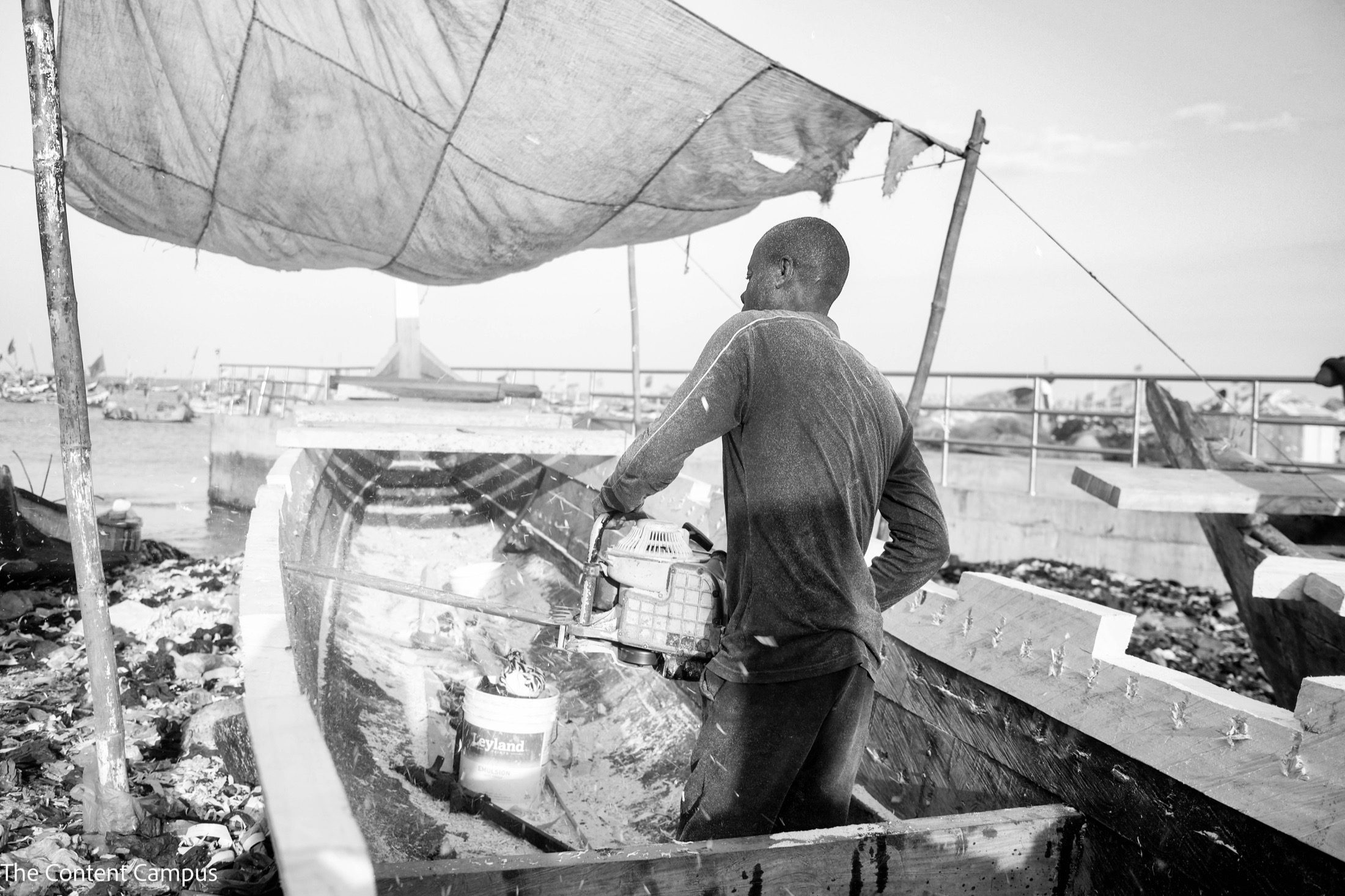In black and white, a man stands inside an unfinished wooden boat and cuts the hull with a motorized saw, under a fabric canopy beside the water.
