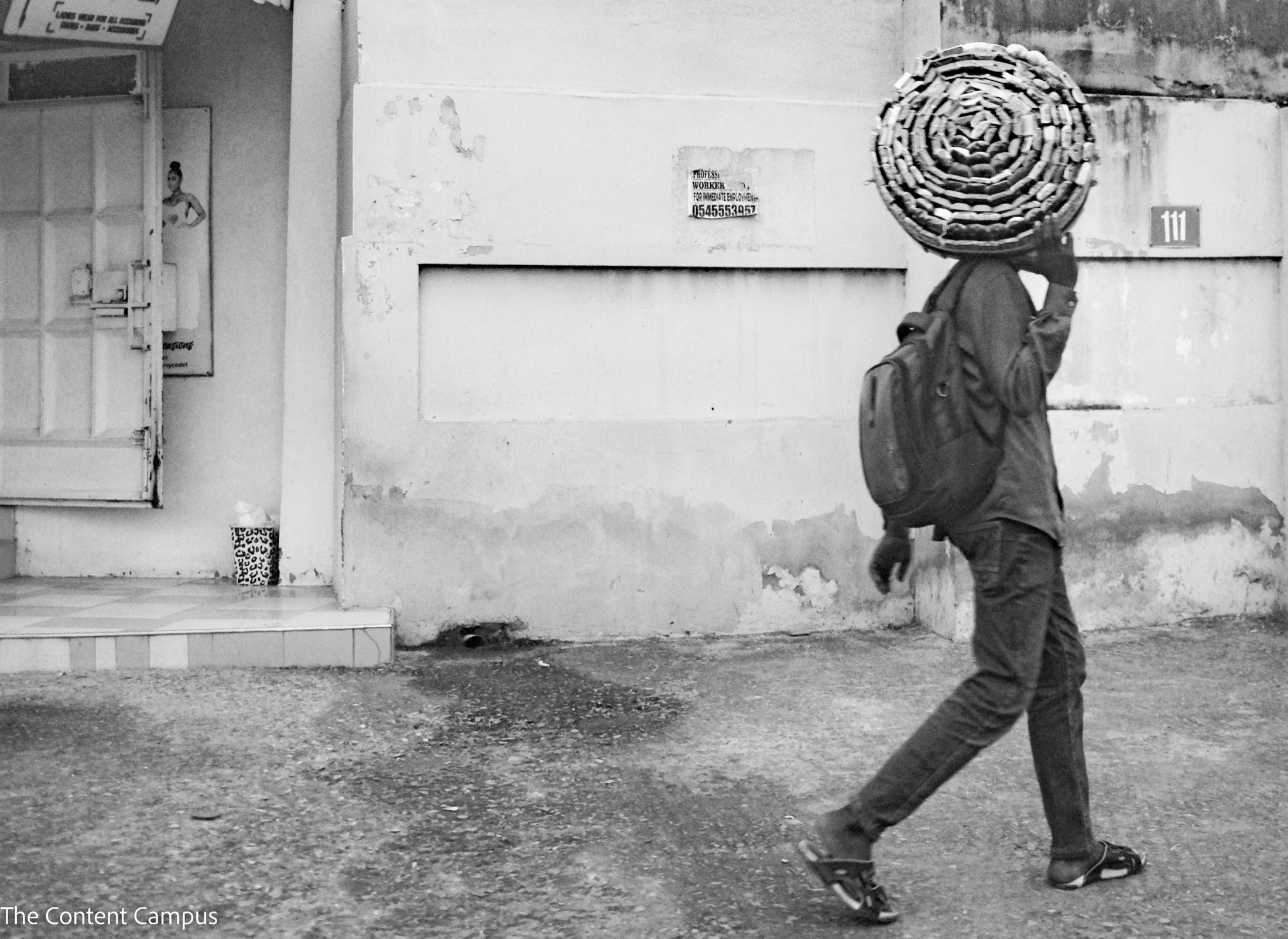 In black and white, a person in a backpack and sandals walks along a street past weathered walls, carrying a large coiled roll on their head.