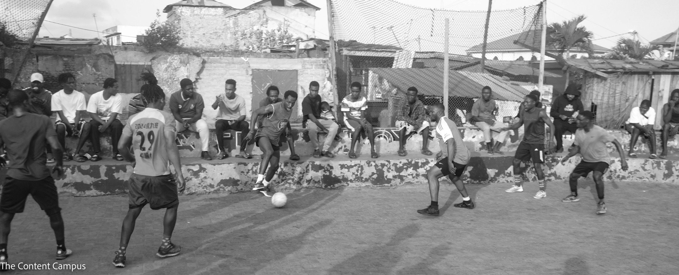 In black and white, men play football on a paved court while a row of spectators sits on a low wall beneath a net, with rooftops behind.