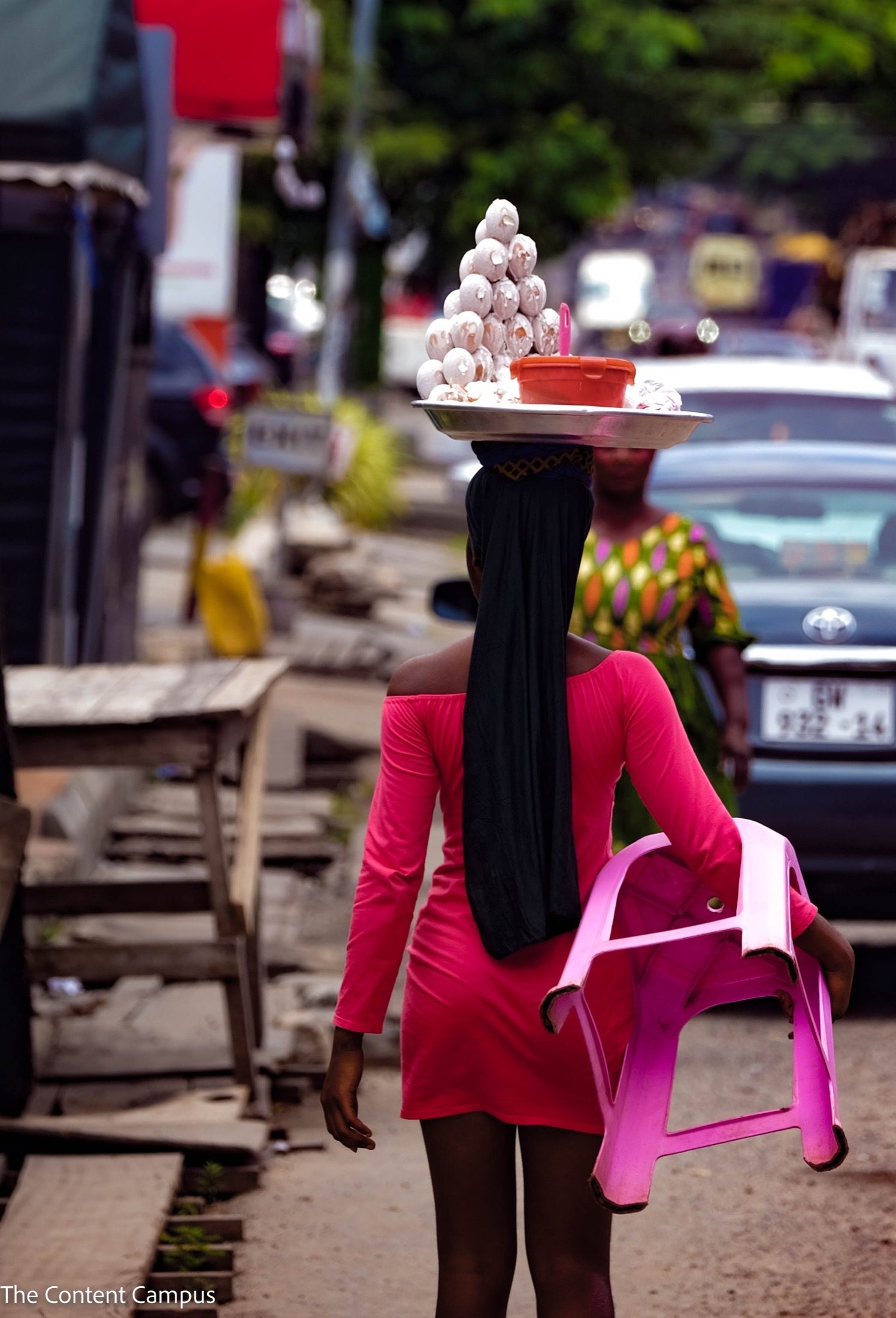 Seen from behind, a woman in a pink dress walks along a roadside, balancing a tray of white food items on her head and carrying a pink plastic chair.