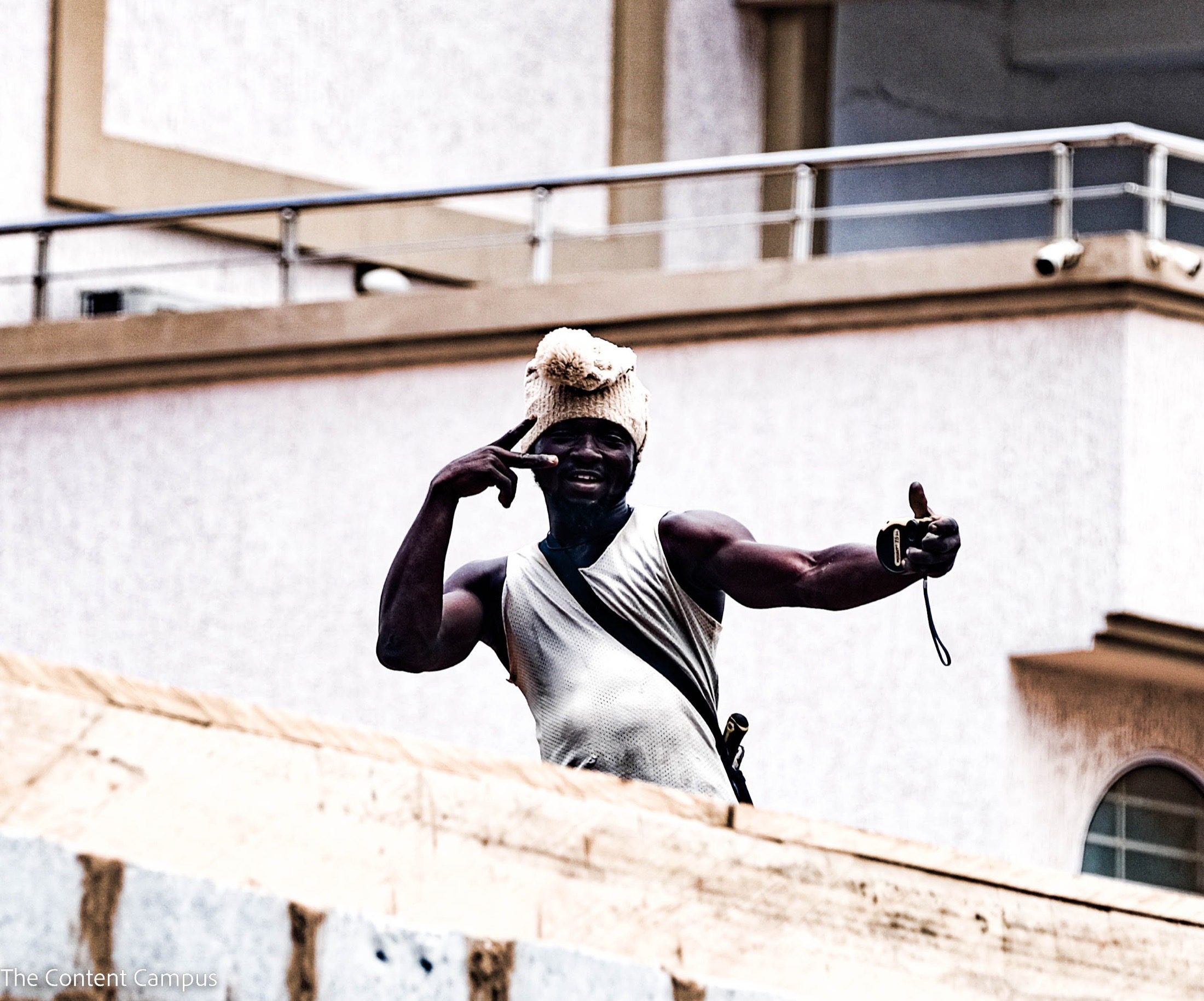 A man in a knit pom-pom hat and sleeveless white top gestures with one hand near his eye and holds a small object in the other, above a rooftop wall.