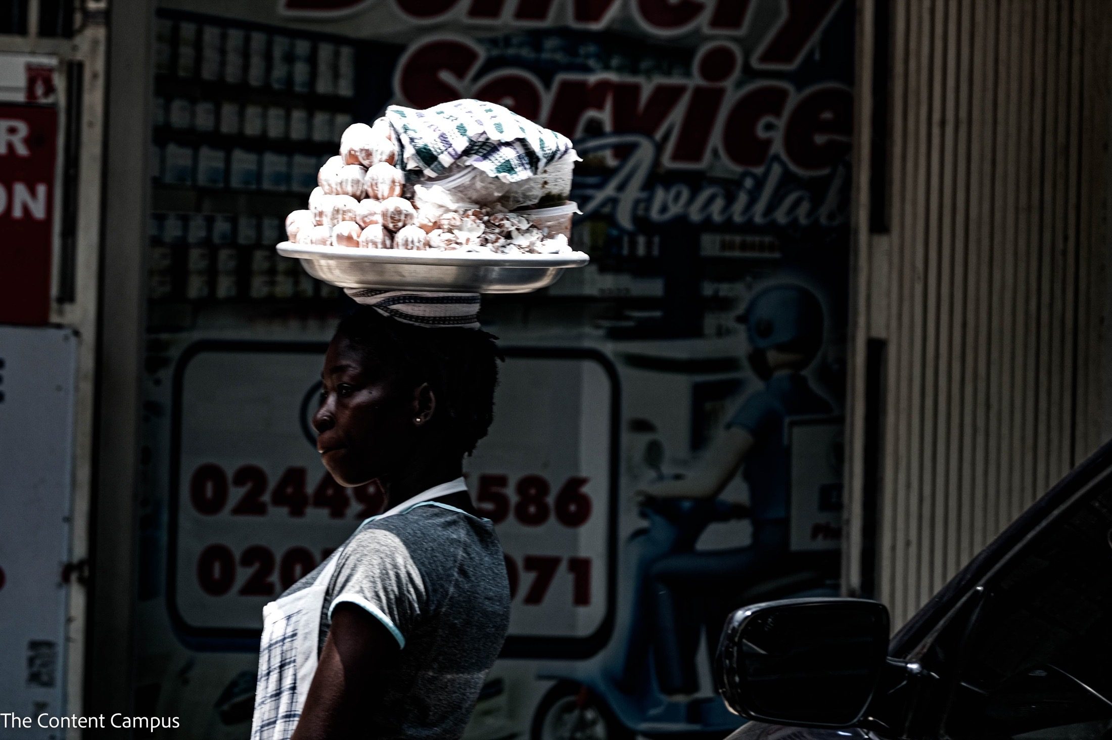 In profile, a woman with a tray of eggs and wrapped food on her head stands in harsh light before a delivery-service sign and parked car.