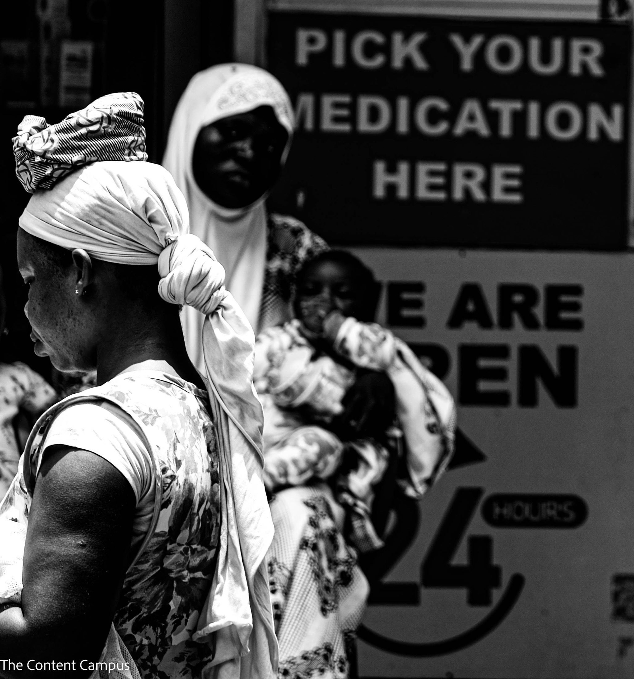 In black and white, a woman in a knotted head wrap looks down in the foreground while a hooded figure and a child stand blurred before signs.