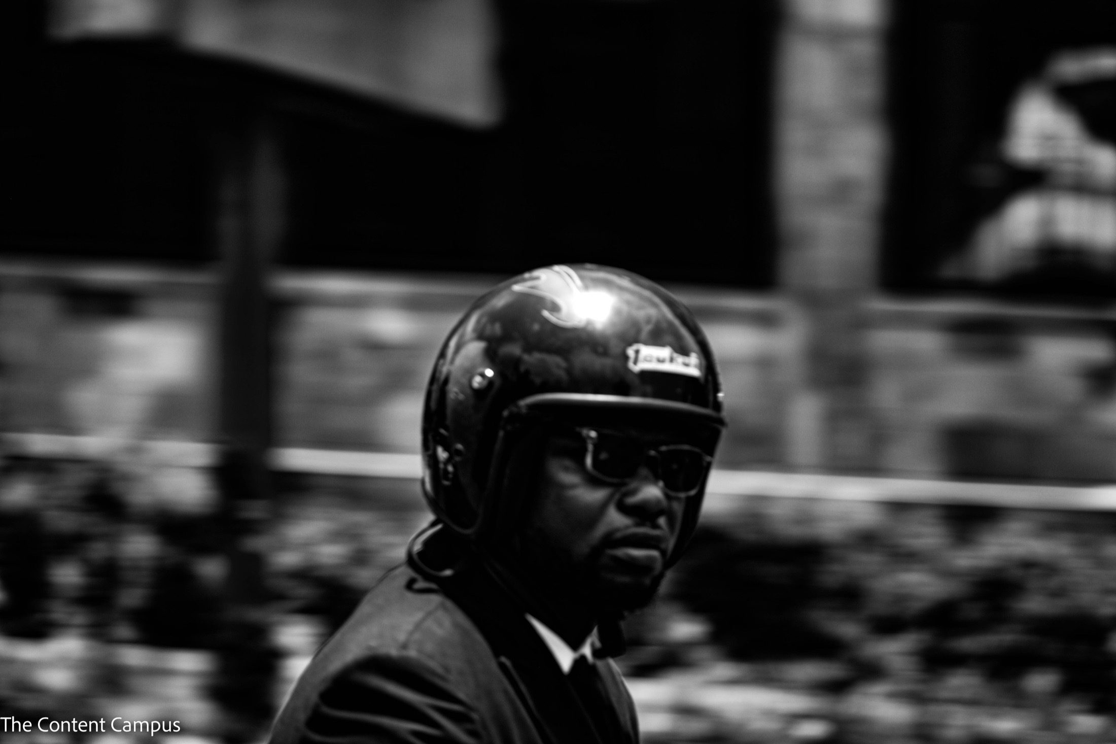 In black and white, a man in a glossy open-face helmet, dark sunglasses and a dark jacket looks toward the camera against a motion-blurred background.