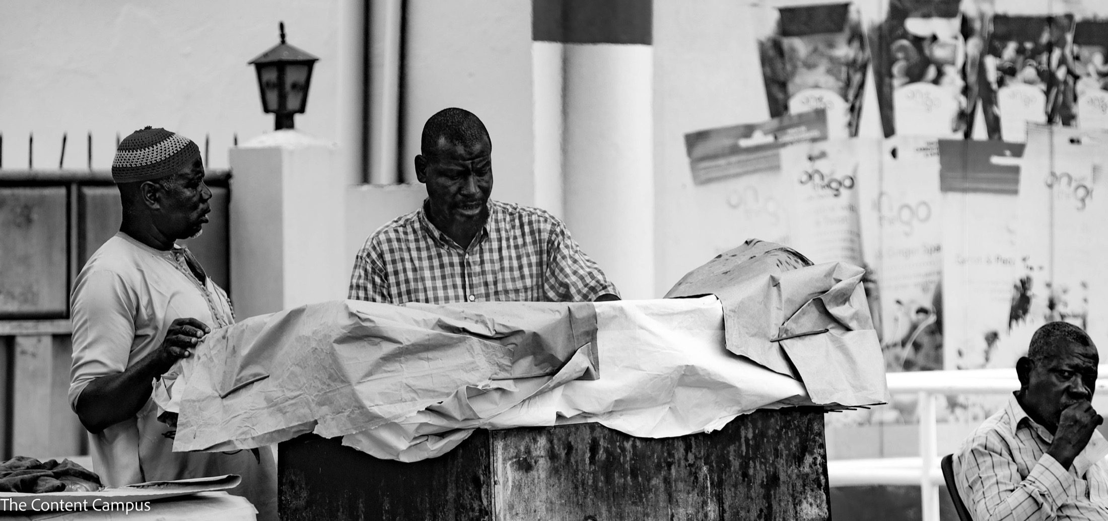 In black and white, two men work and talk behind a rusty counter covered with crumpled paper while a third man sits at right, hand to his mouth.