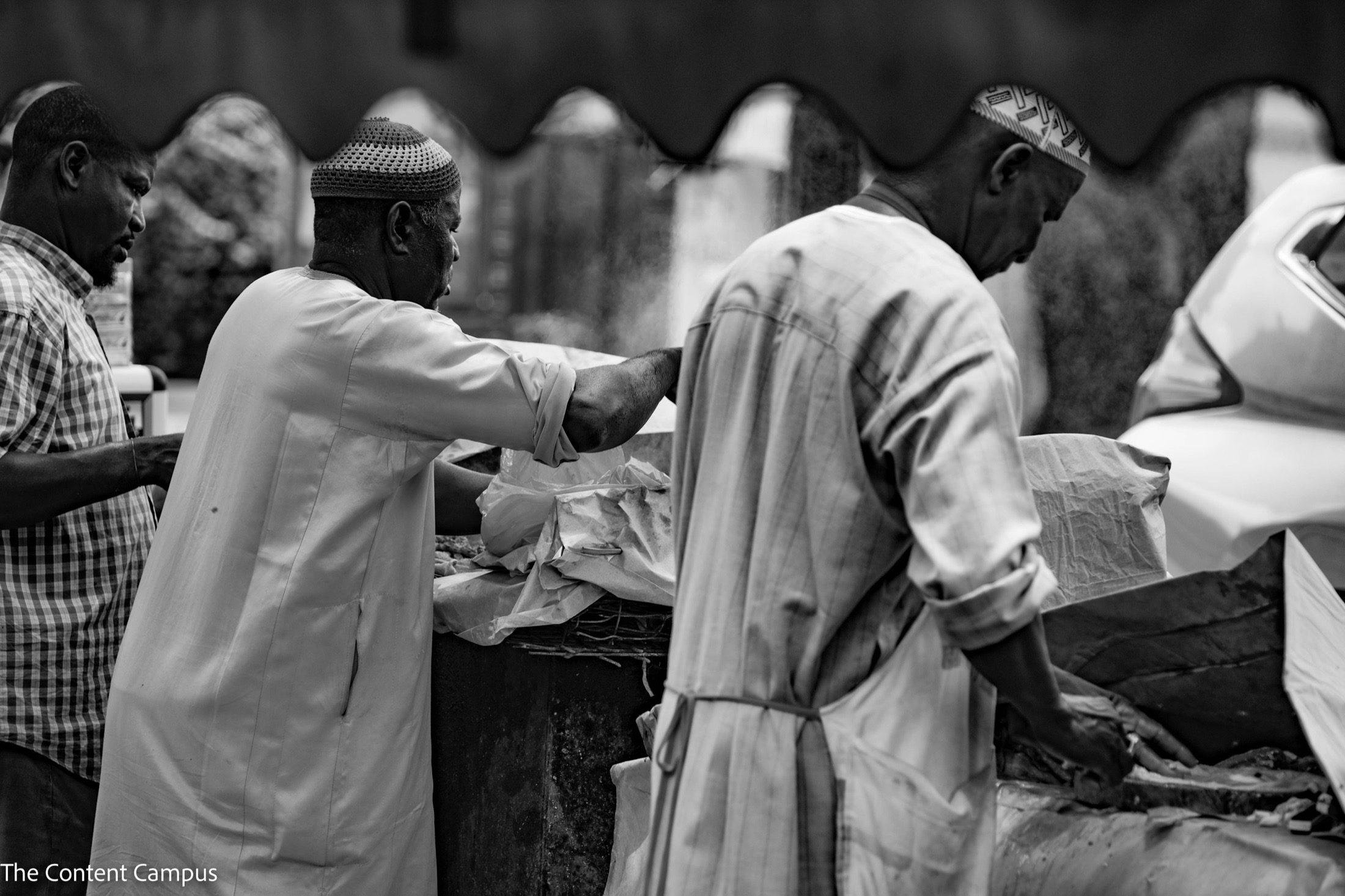 In black and white, two men in long light tunics and caps work at an outdoor stall with paper-wrapped goods, while a third man in a checked shirt watches from the left.