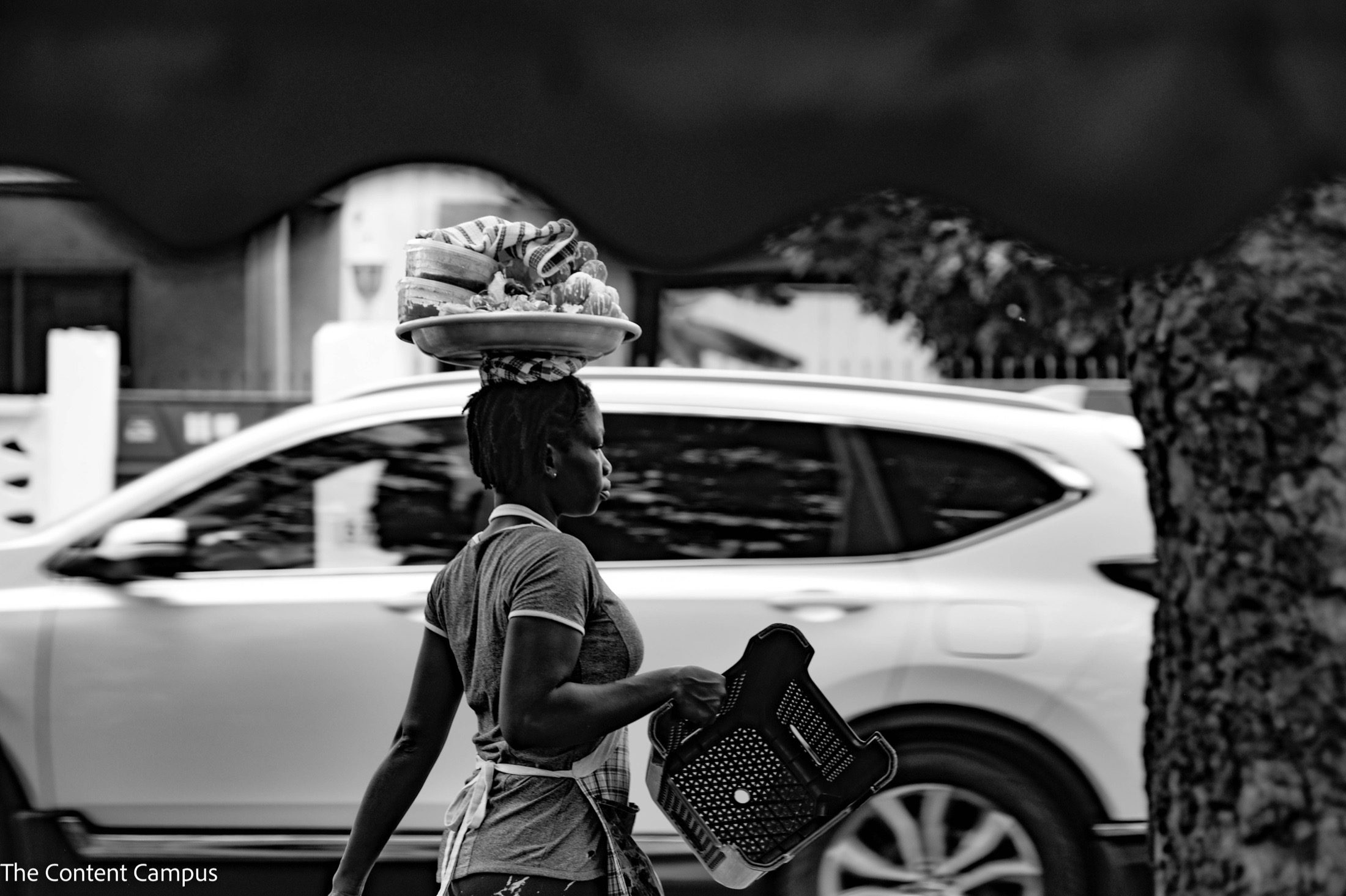 In black and white, a woman in profile walks past a blurred white SUV, balancing a tray of goods on her head and carrying an upturned plastic crate.