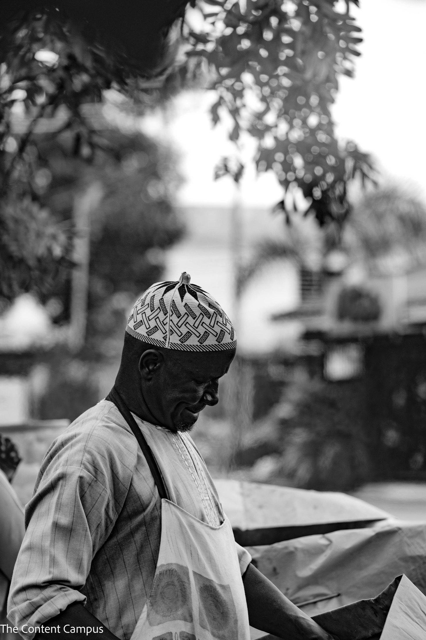 In black and white, a man in a patterned cap and apron looks down while working at an outdoor stall, with trees and buildings softly blurred behind him.