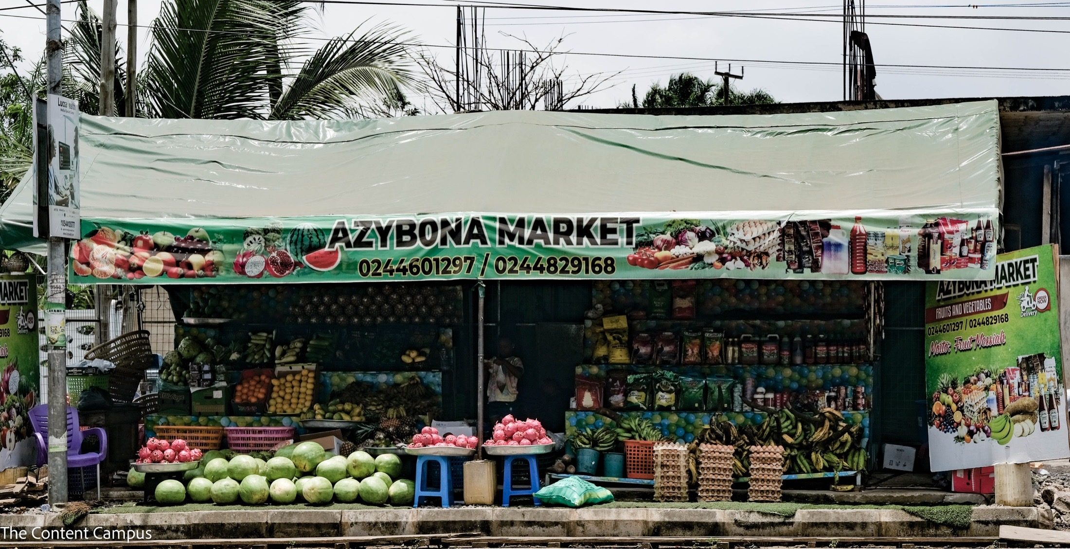 A roadside market stall under a green tarpaulin banner reading "Azybona Market" displays watermelons, dragon fruit, plantains, egg crates, bottled goods and packaged groceries on overcast daylight.