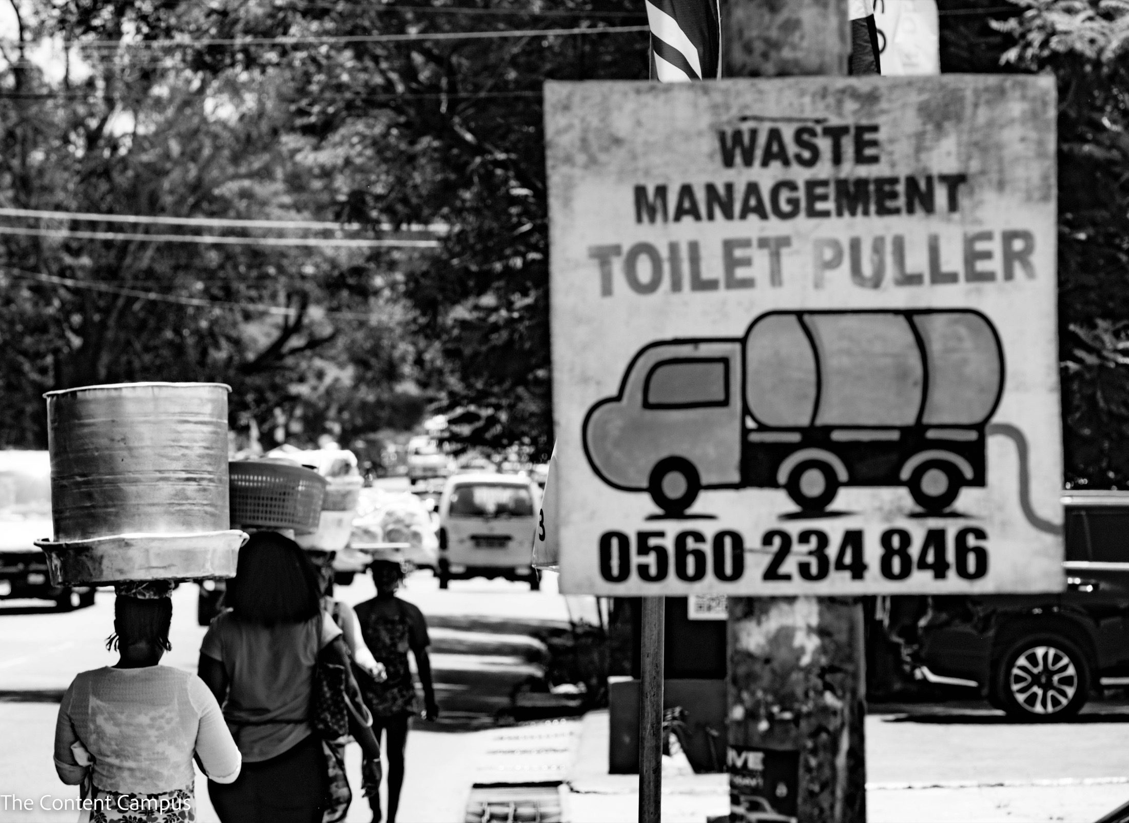 In black and white, women walk away along a tree-lined street carrying large metal basins and baskets on their heads, beside a sign reading "Waste Management Toilet Puller" with a tanker truck drawing.