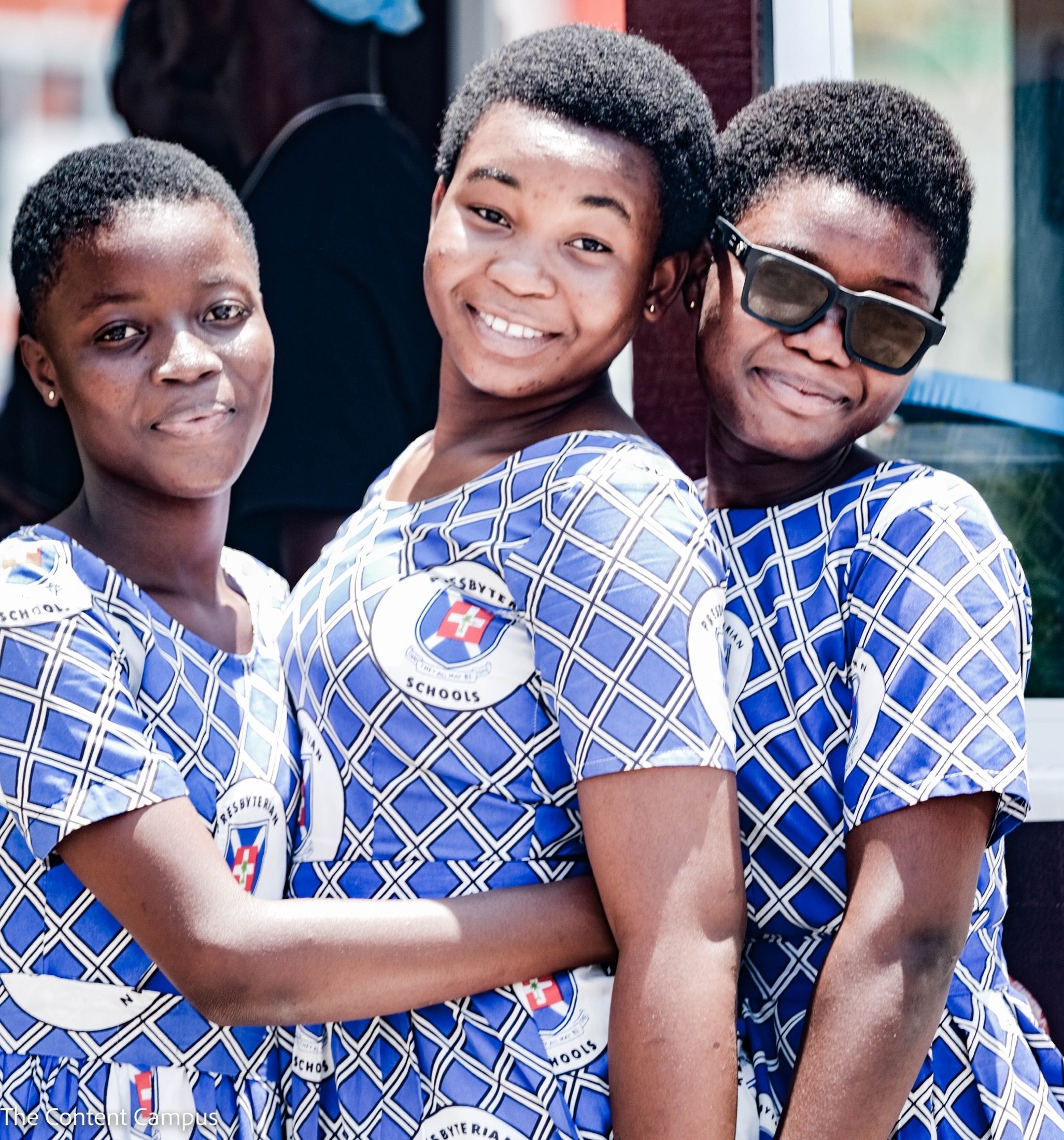 Three young women with short hair, wearing matching blue-and-white patterned school dresses marked "Presbyterian Schools," lean together and smile at the camera in bright sunlight; one wears dark sunglasses.