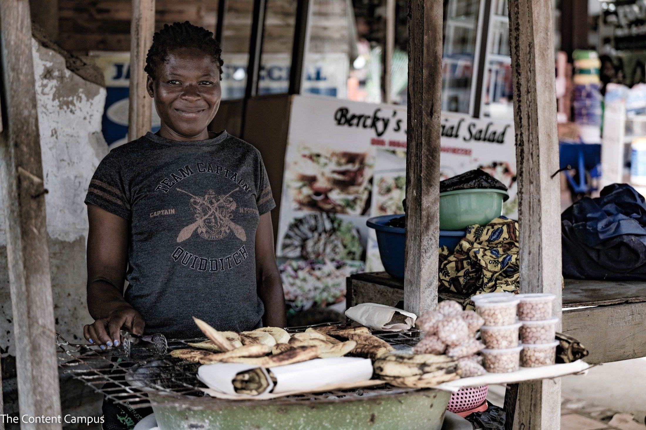 A woman in a grey graphic T-shirt stands behind a street stall grill of roasted plantains, with cups of peanuts beside her and a food sign behind.