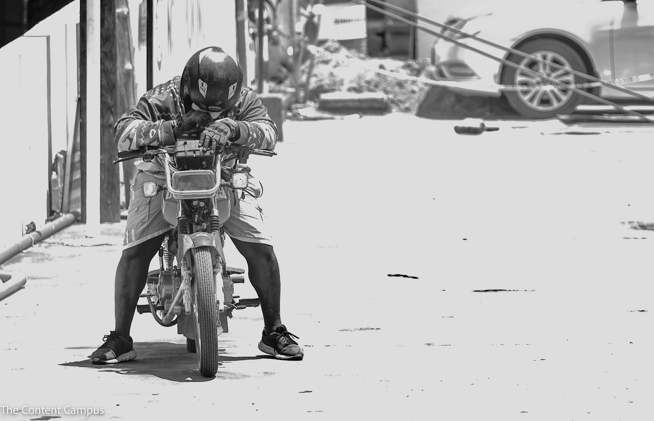 In harsh, high-contrast black and white, a helmeted rider in shorts and sneakers leans forward on parked motorbike handlebars on a sunlit street, a car behind.