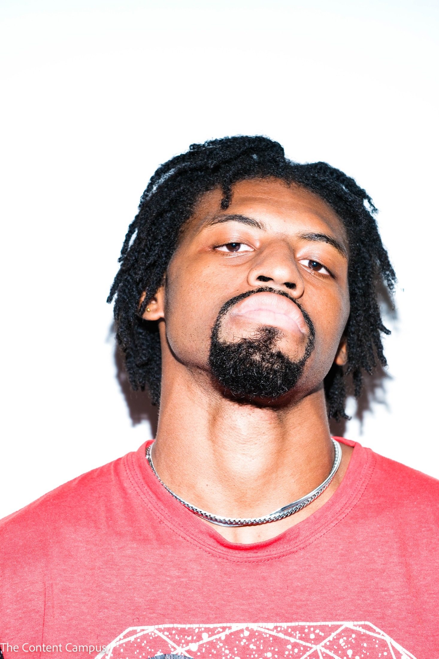 A man with twisted hair and a goatee tilts his head back, chin raised, wearing a red t-shirt and silver chain under harsh, bright flash.