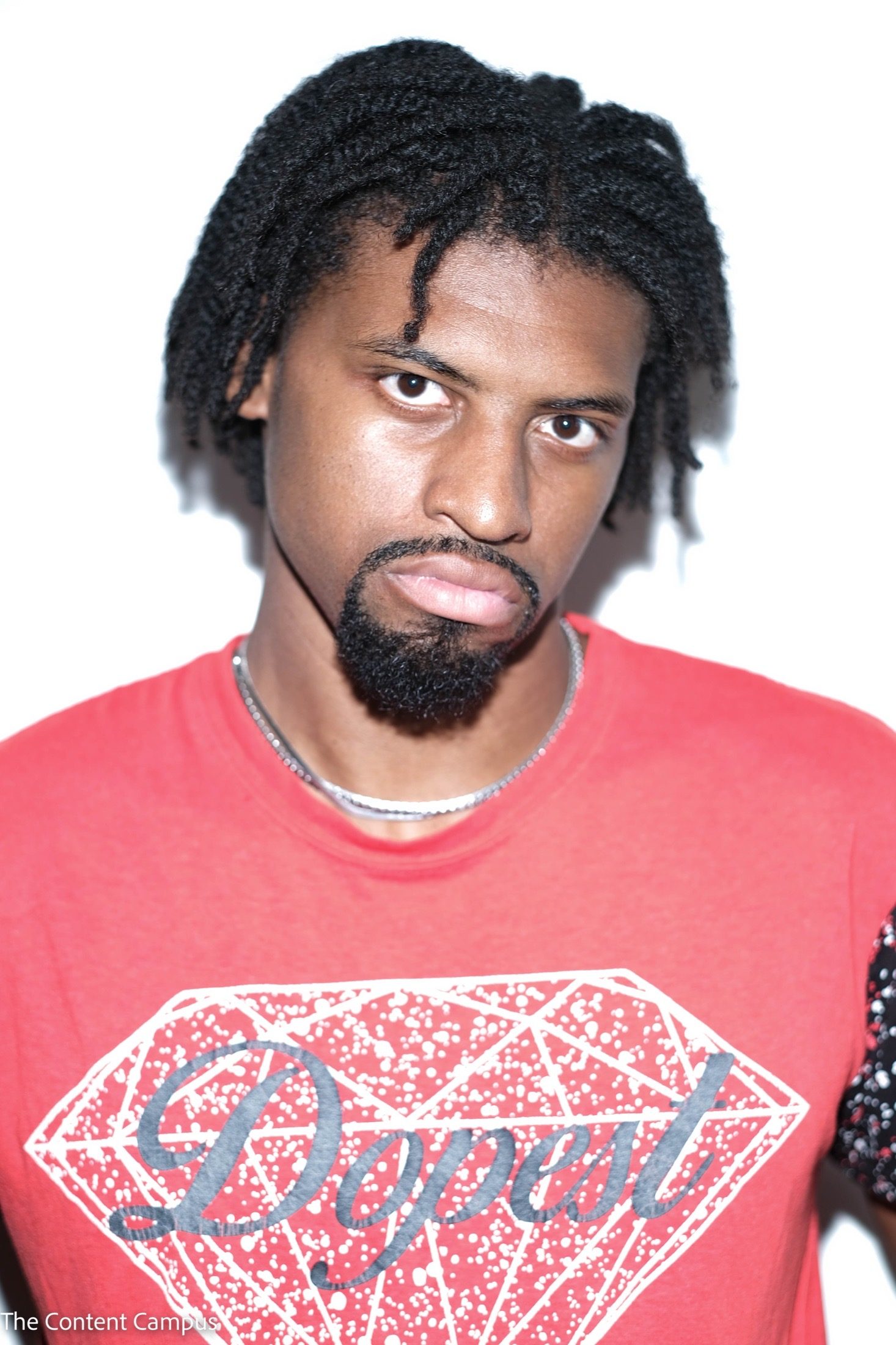 A man with twisted hair and a goatee looks at the camera, wearing a red graphic t-shirt with a diamond print and a silver chain.
