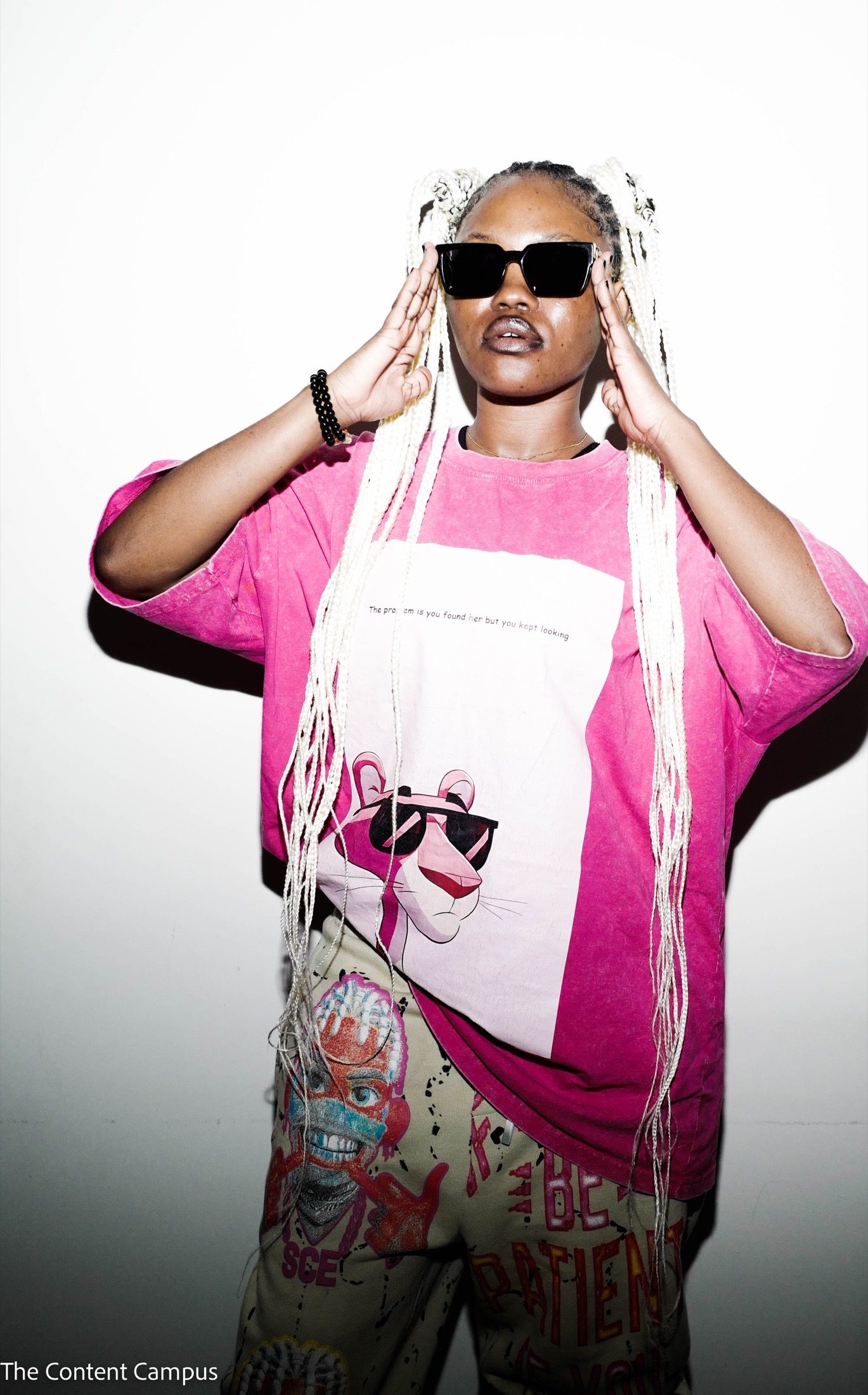 A person with long white braids and square black sunglasses raises both hands to their face, wearing a pink graphic T-shirt and patterned trousers against a bright white wall.