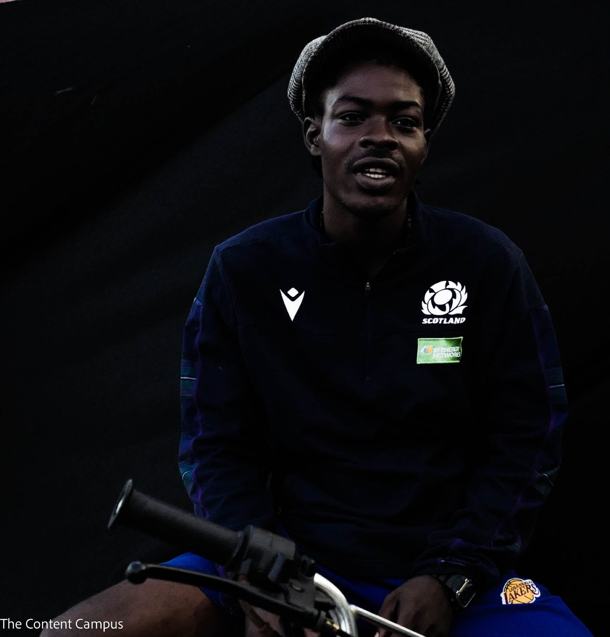 A person in a tweed flat cap and a navy Scotland team jacket sits behind bicycle handlebars, speaking to the camera against a dark background.