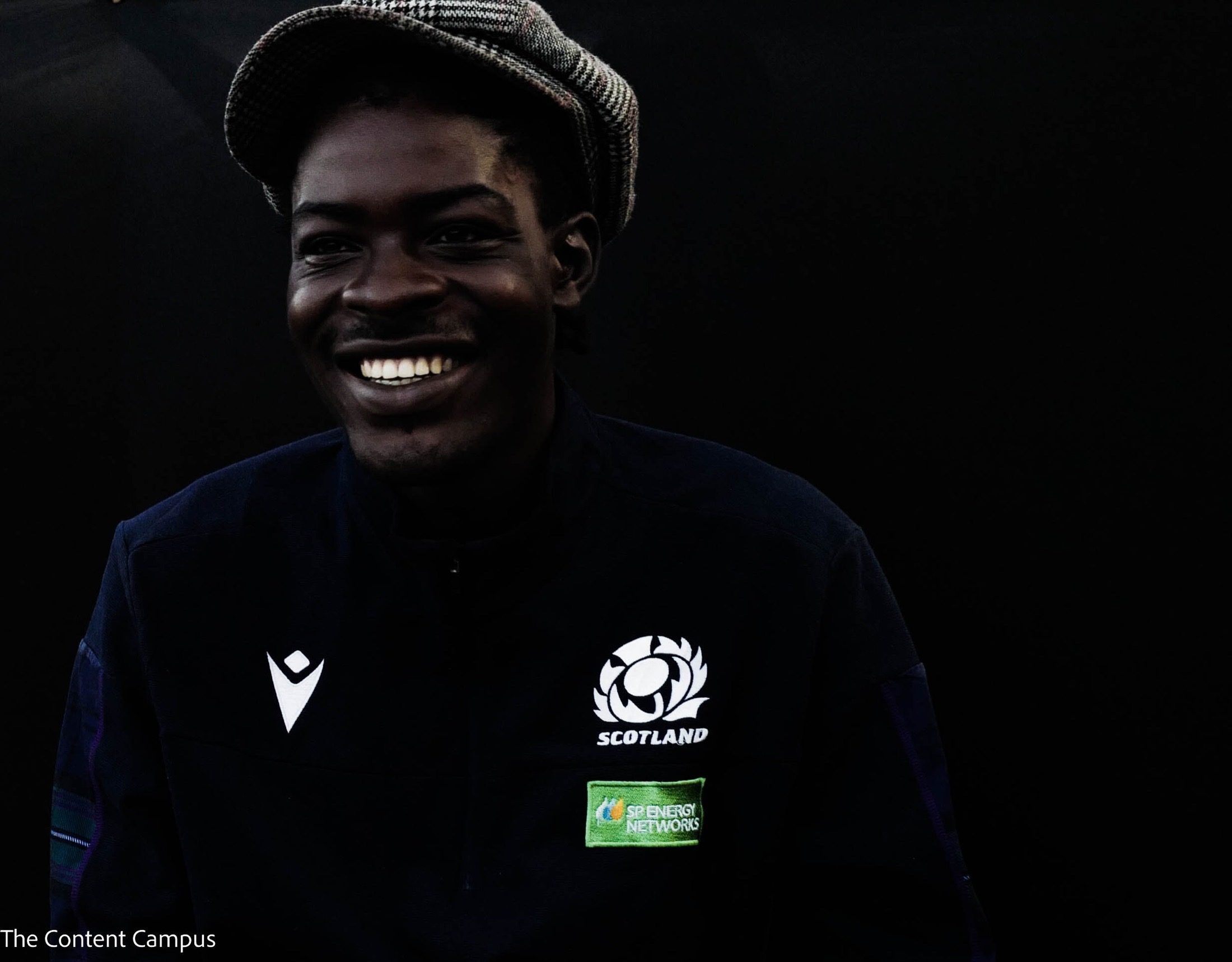 A smiling person in a checked flat cap and a navy Scotland jacket with sponsor patch, lit against a near-black background.