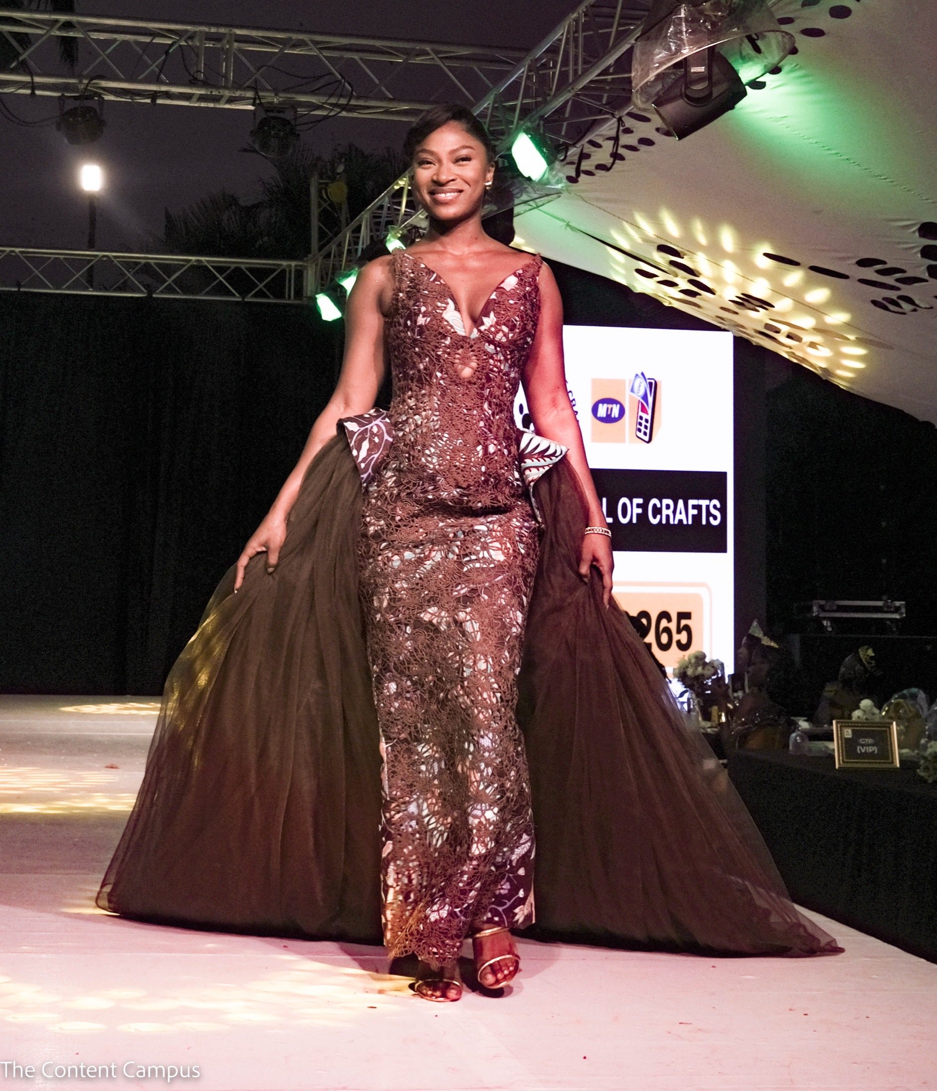 At night, a smiling woman walks a lit runway in a brown lace gown with a sheer train, holding the fabric at her sides.