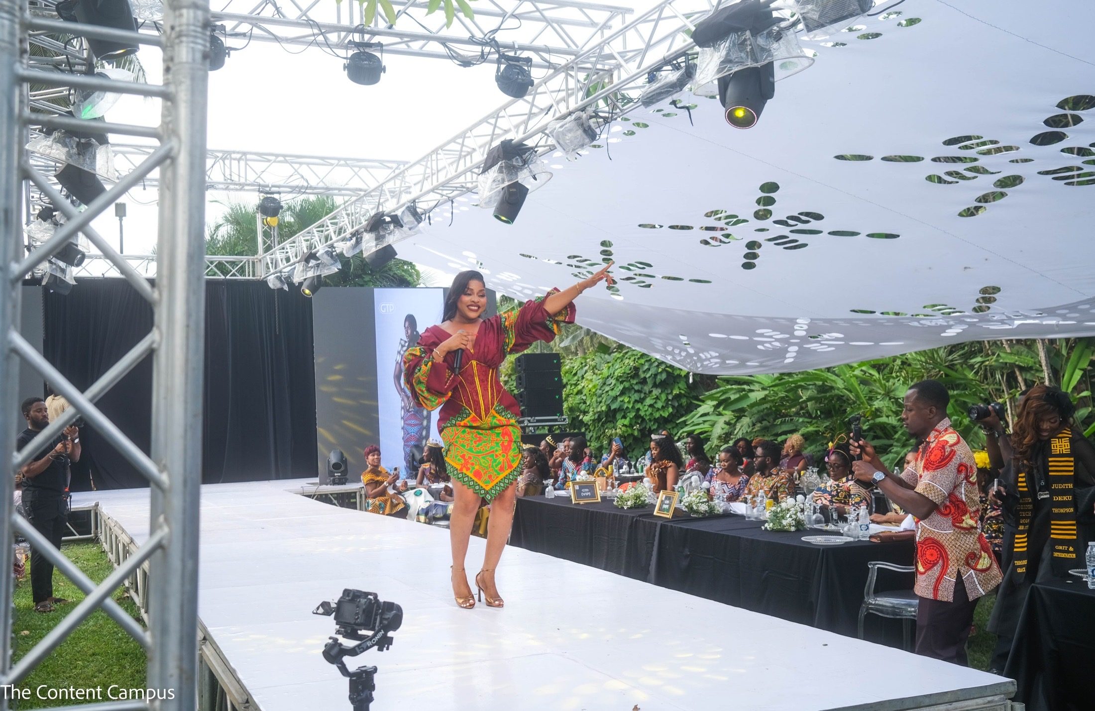 On a white outdoor runway beneath a perforated canopy, a woman in a patterned maroon and green dress points upward while holding a microphone; guests and camera operators look on.