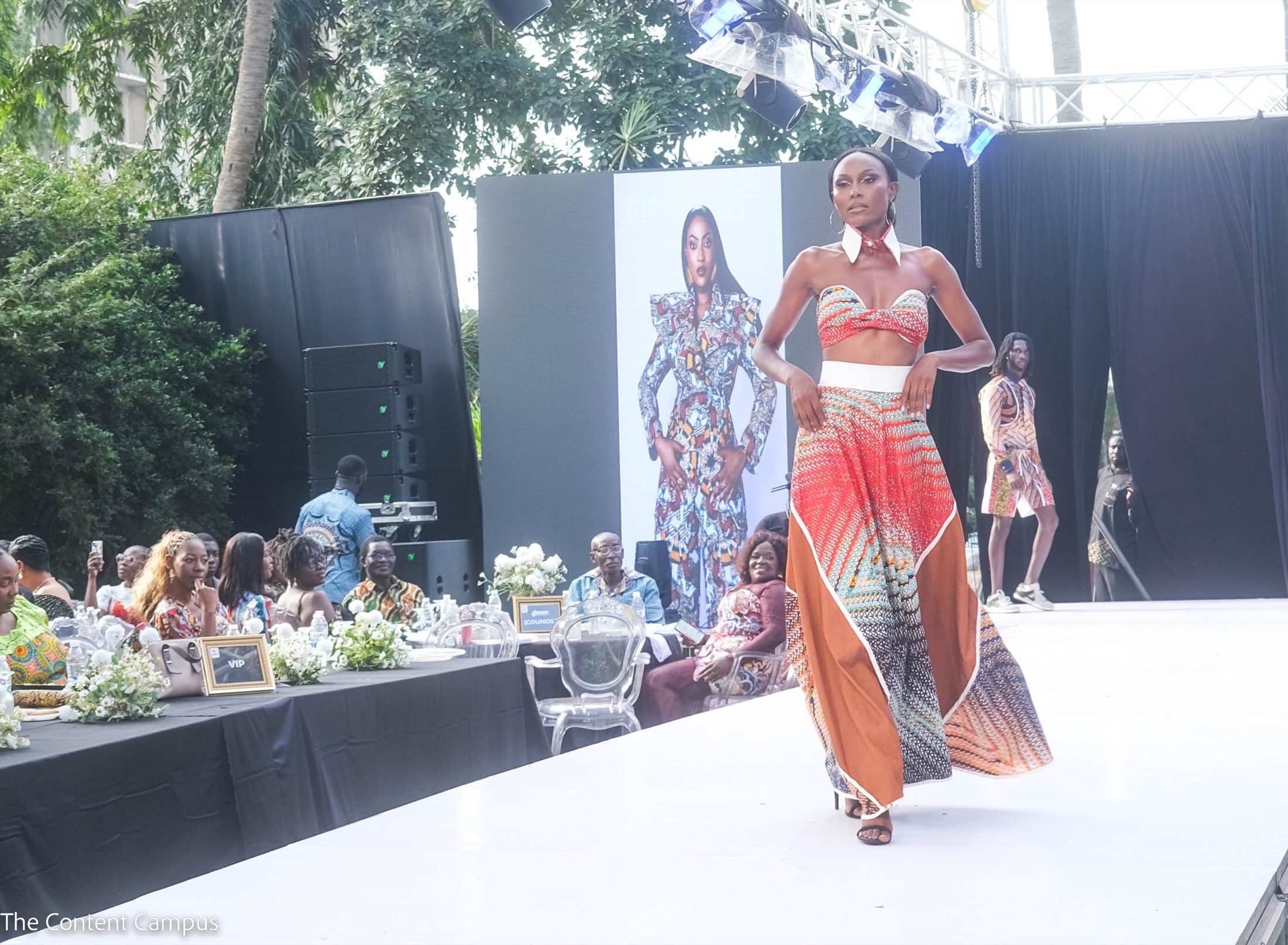 A model in a patterned crop top, white collar and wide asymmetric skirt walks a runway in daylight as seated guests and another model look on.