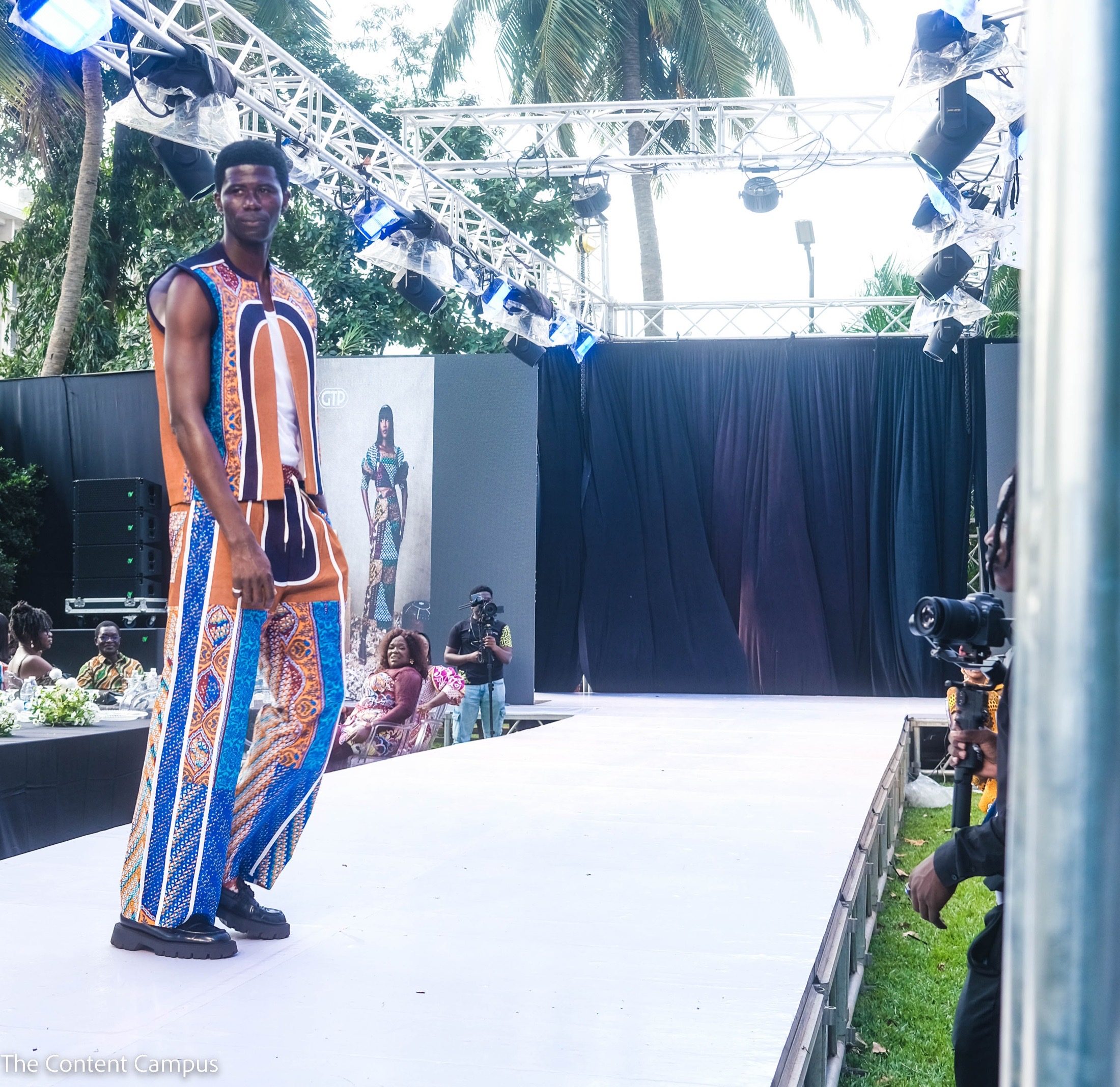A man in a sleeveless orange, navy and blue patterned vest and matching trousers walks a white runway as camera operators film beneath stage lighting rigs.