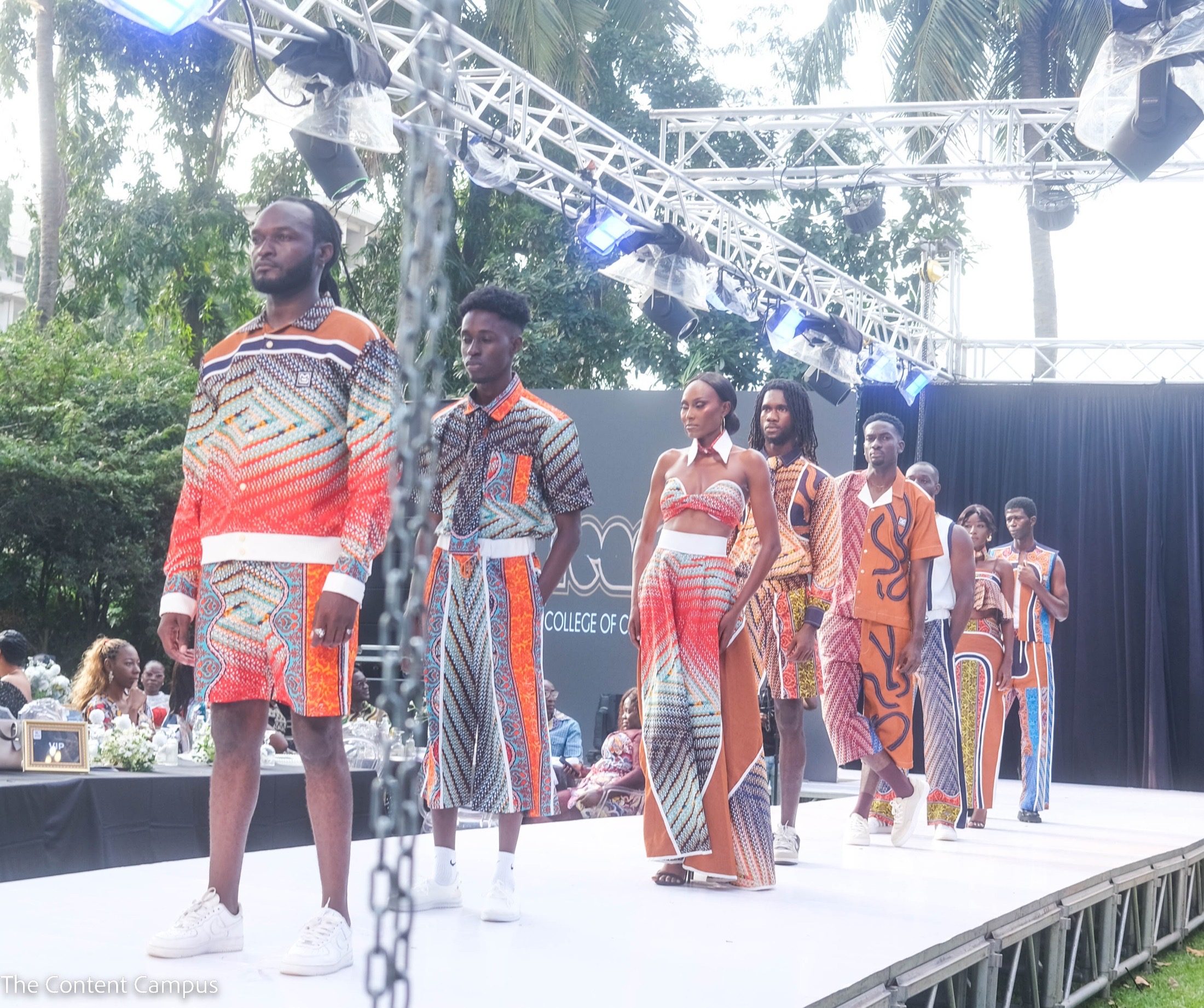 A line of models in coordinated orange, blue and white patterned outfits stands and walks along a white outdoor runway beneath trees and lighting trusses.